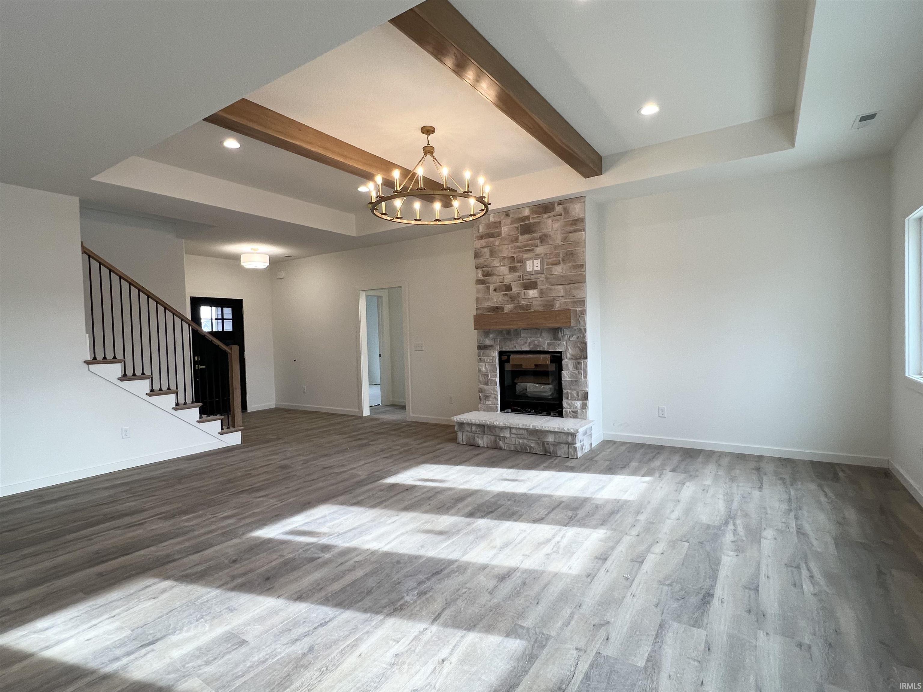 Unfurnished living room featuring beamed ceiling, a stone fireplace, light wood-style floors, stairway, and a chandelier