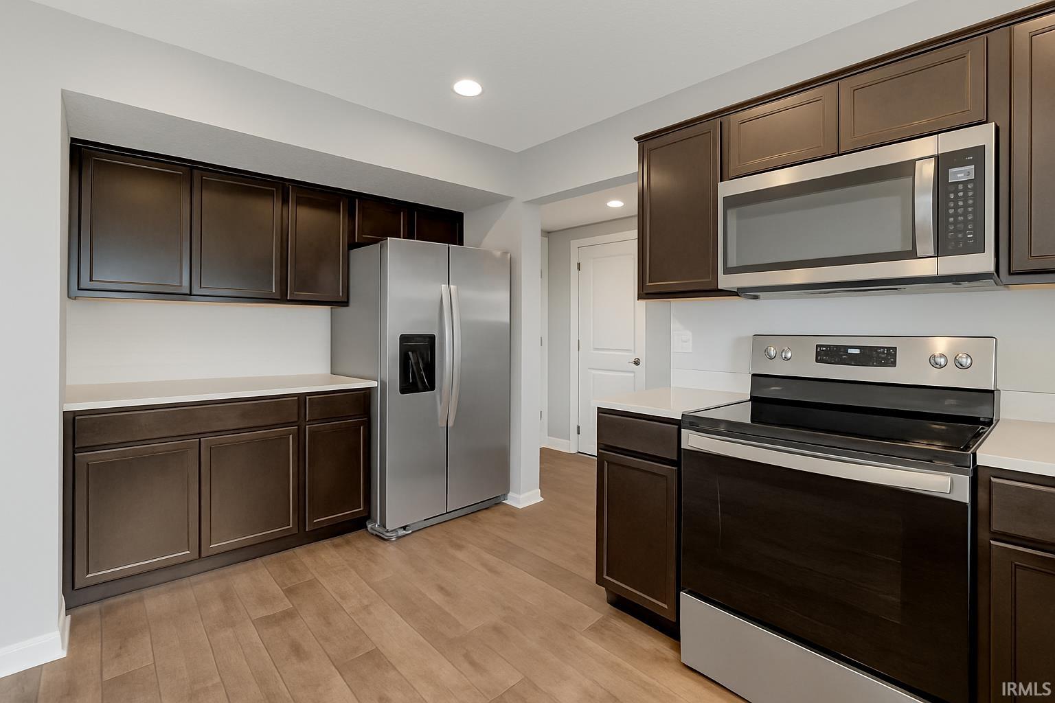 Kitchen with appliances with stainless steel finishes, dark brown cabinetry, light countertops, light wood-style flooring, and recessed lighting