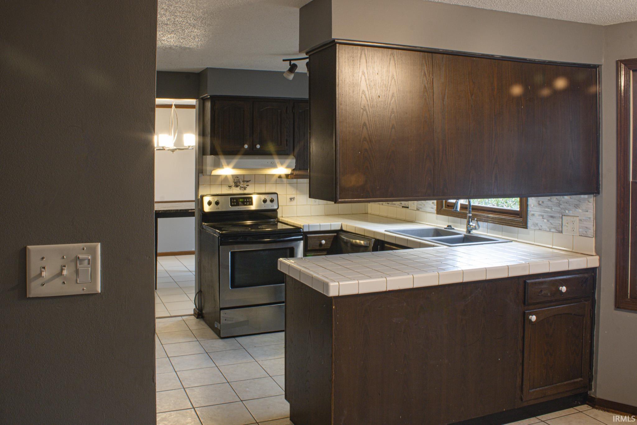 Kitchen with dark brown cabinets, light tile patterned floors, stainless steel range with electric stovetop, tile countertops, and decorative backsplash