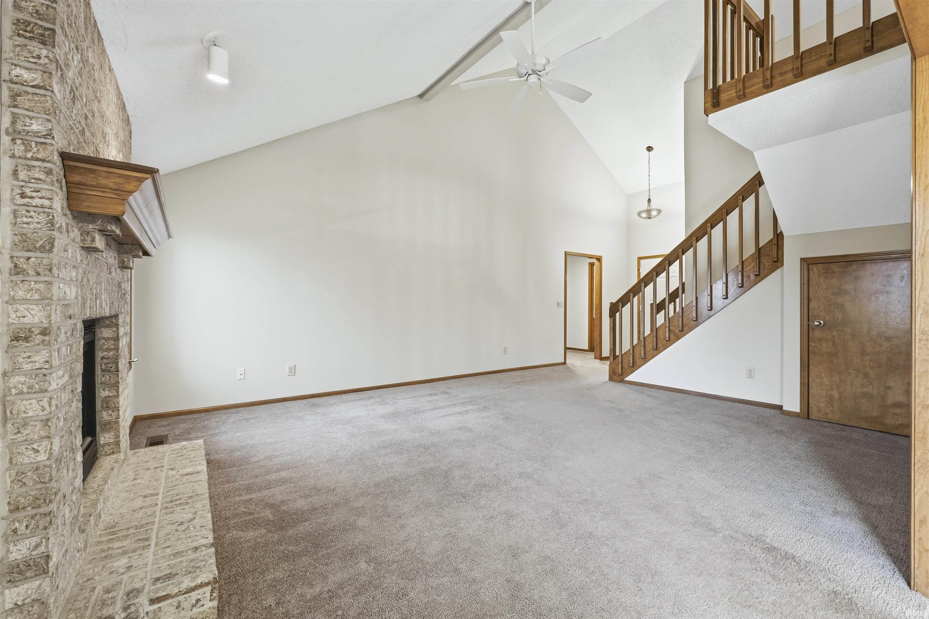 Unfurnished living room featuring stairs, high vaulted ceiling, a brick fireplace, carpet, and a ceiling fan