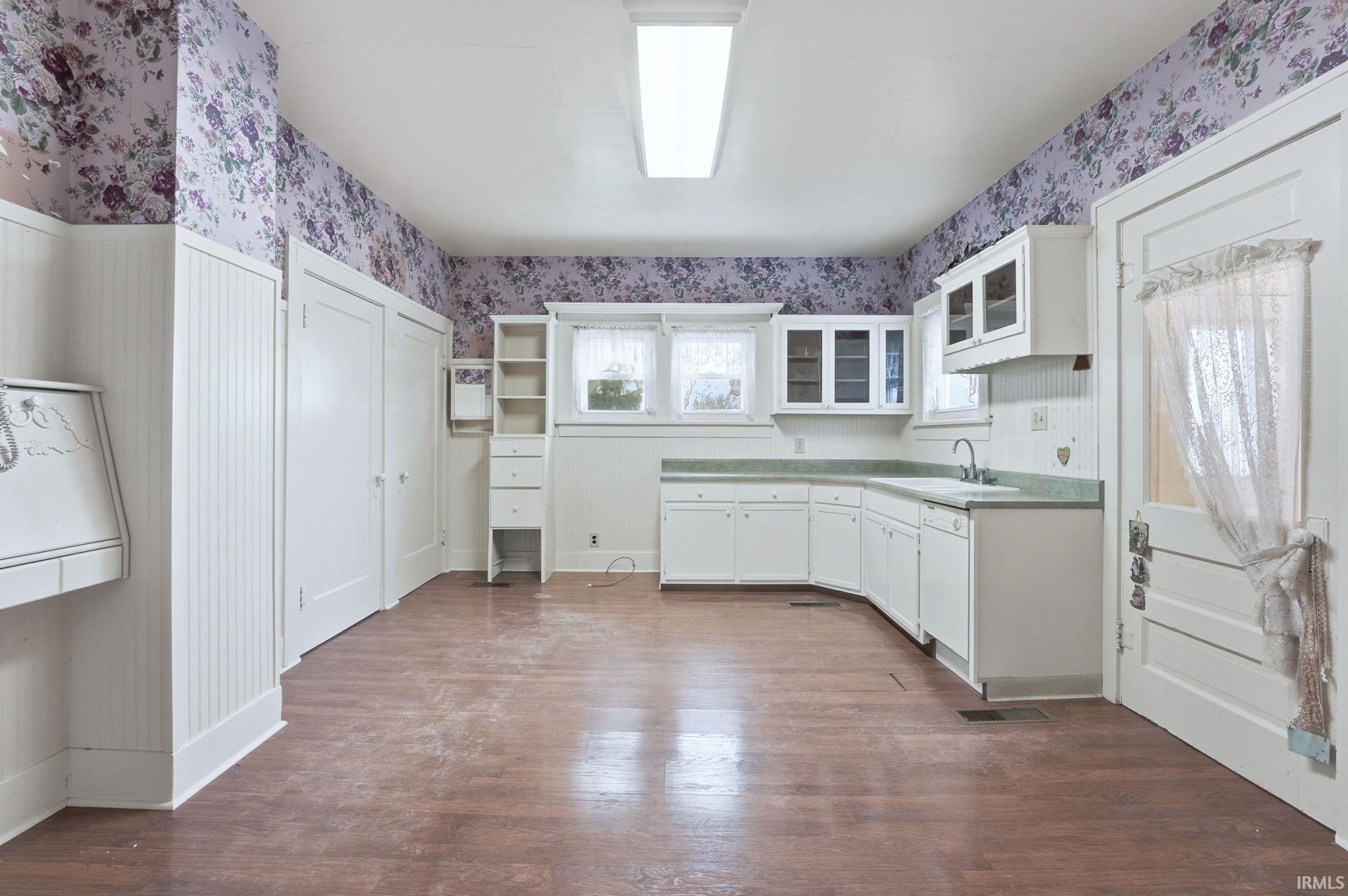 Kitchen with wallpapered walls, white cabinetry, dark wood-type flooring, light countertops, and glass insert cabinets