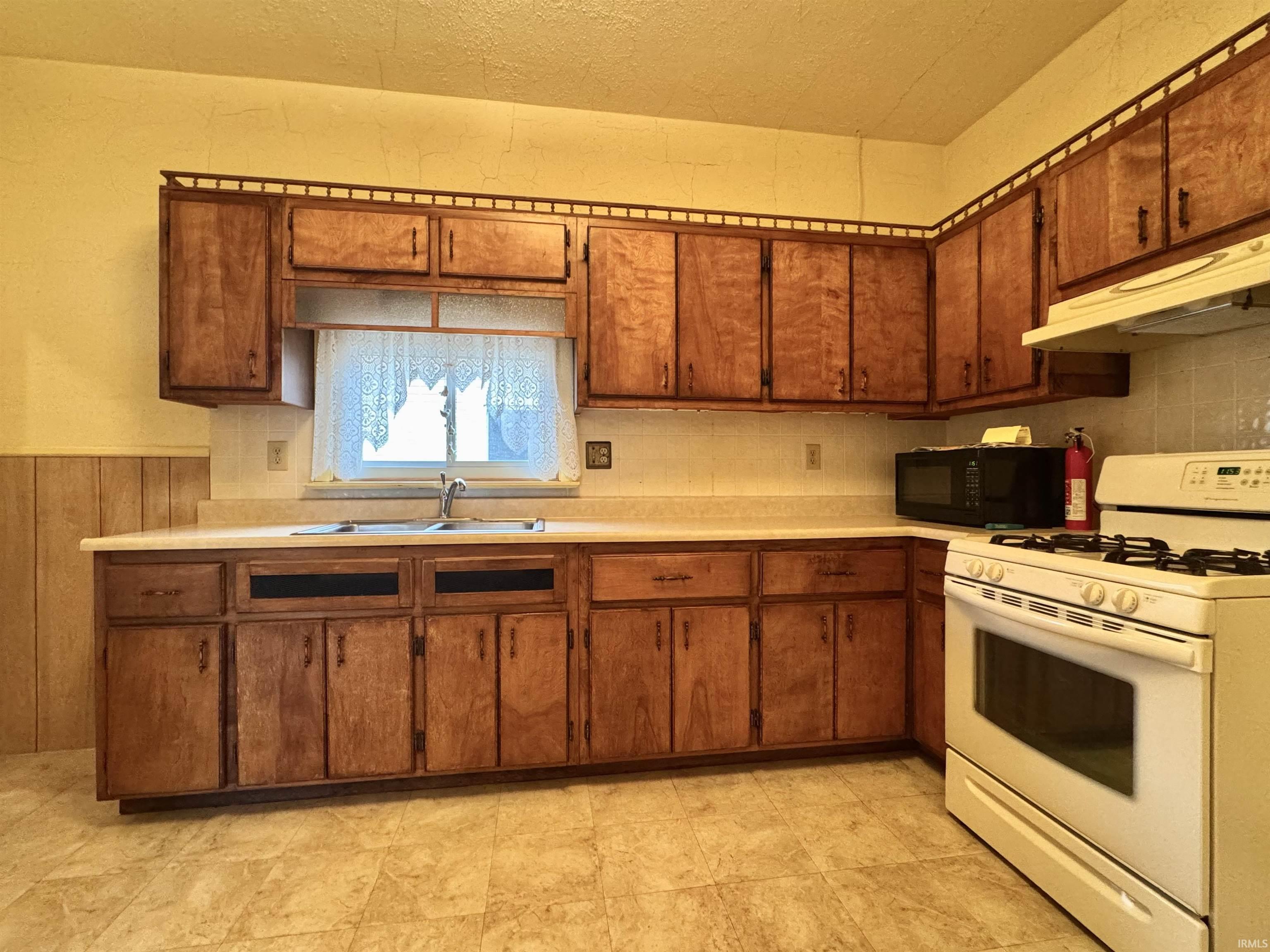 Kitchen with white range with gas stovetop, light countertops, under cabinet range hood, black microwave, and brown cabinetry