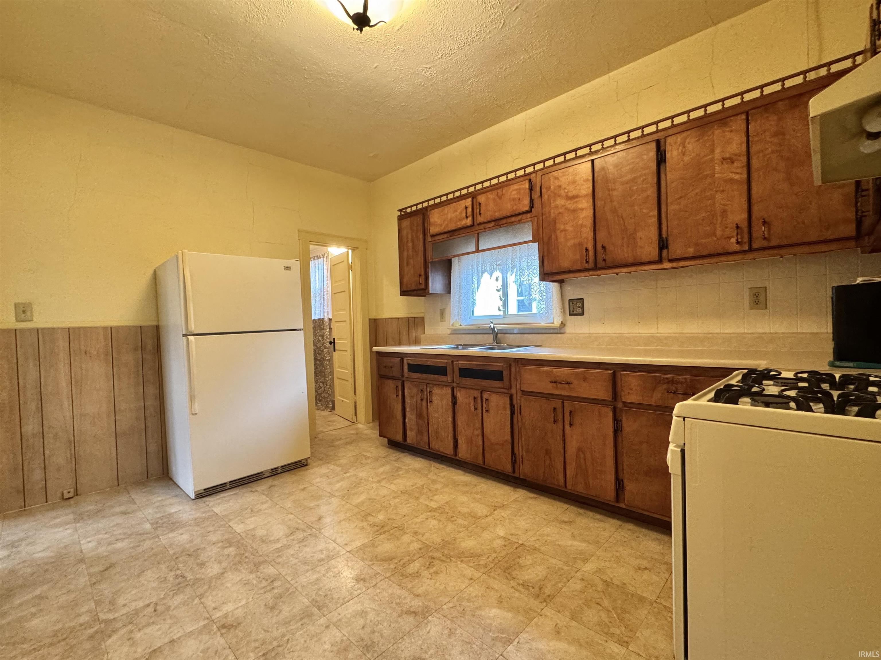 Kitchen with light countertops, white appliances, a textured ceiling, brown cabinets, and wainscoting