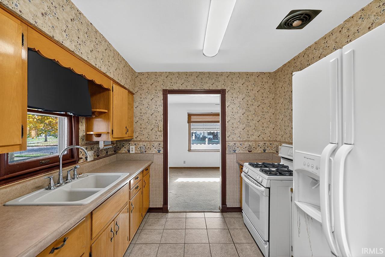 Kitchen featuring white appliances, wallpapered walls, light countertops, light tile patterned floors, and brown cabinetry