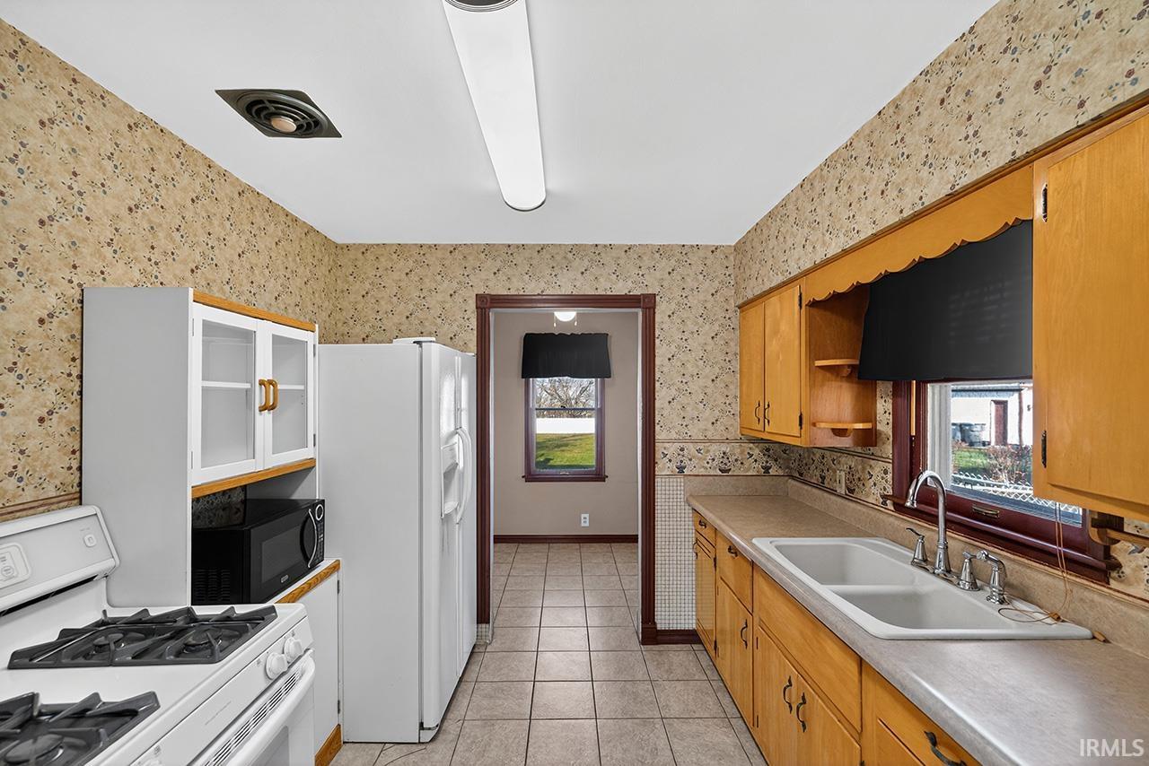 Kitchen featuring wallpapered walls, white appliances, light countertops, light tile patterned floors, and glass insert cabinets