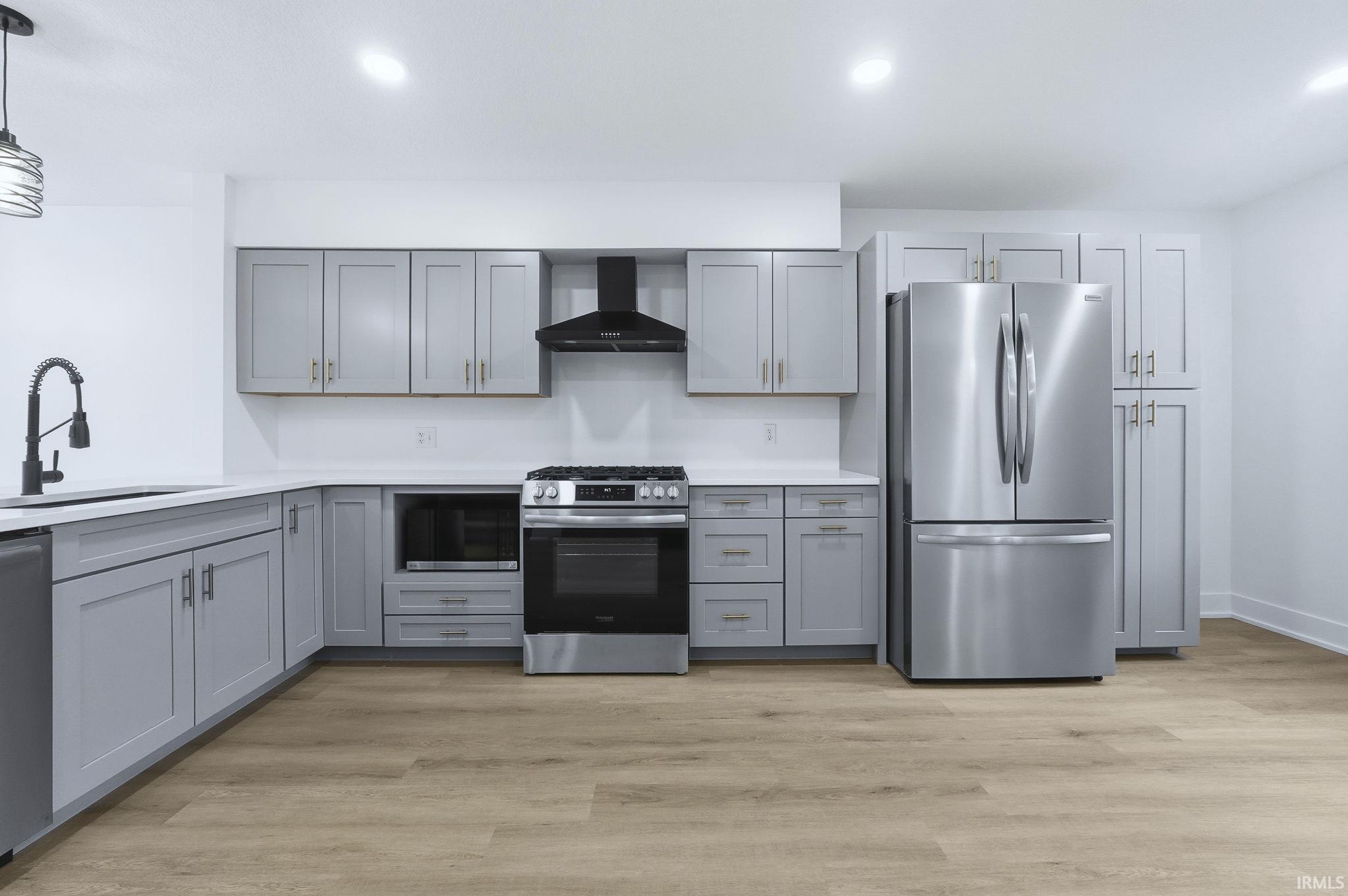 Kitchen with gray cabinetry, appliances with stainless steel finishes, wall chimney exhaust hood, light wood finished floors, and recessed lighting