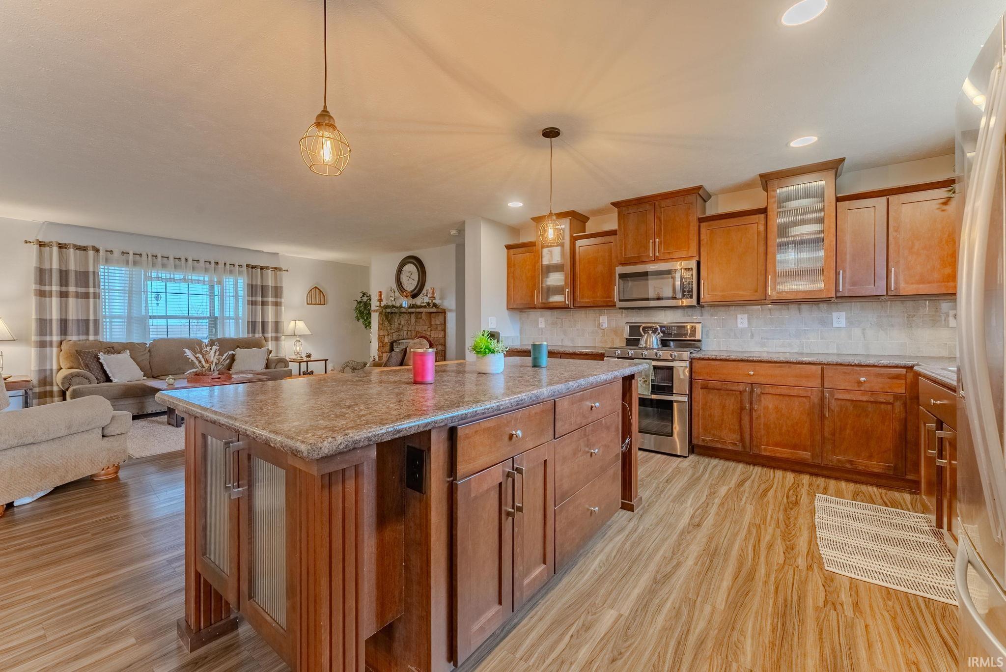 Kitchen with glass insert cabinets, brown cabinetry, hanging light fixtures, stainless steel appliances, and a center island