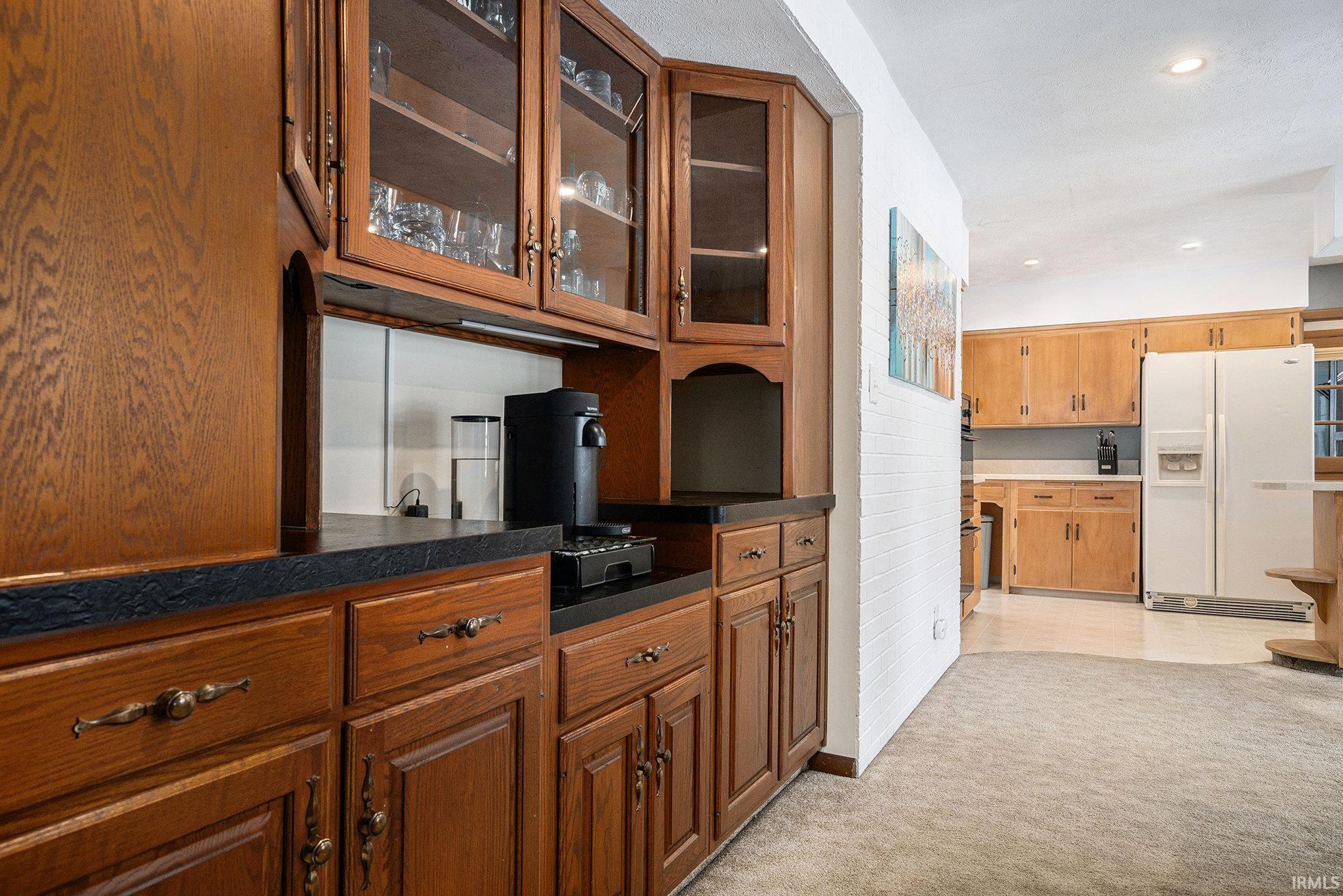 Bar area with dark countertops, glass insert cabinets, white fridge with ice dispenser, brown cabinets, and recessed lighting