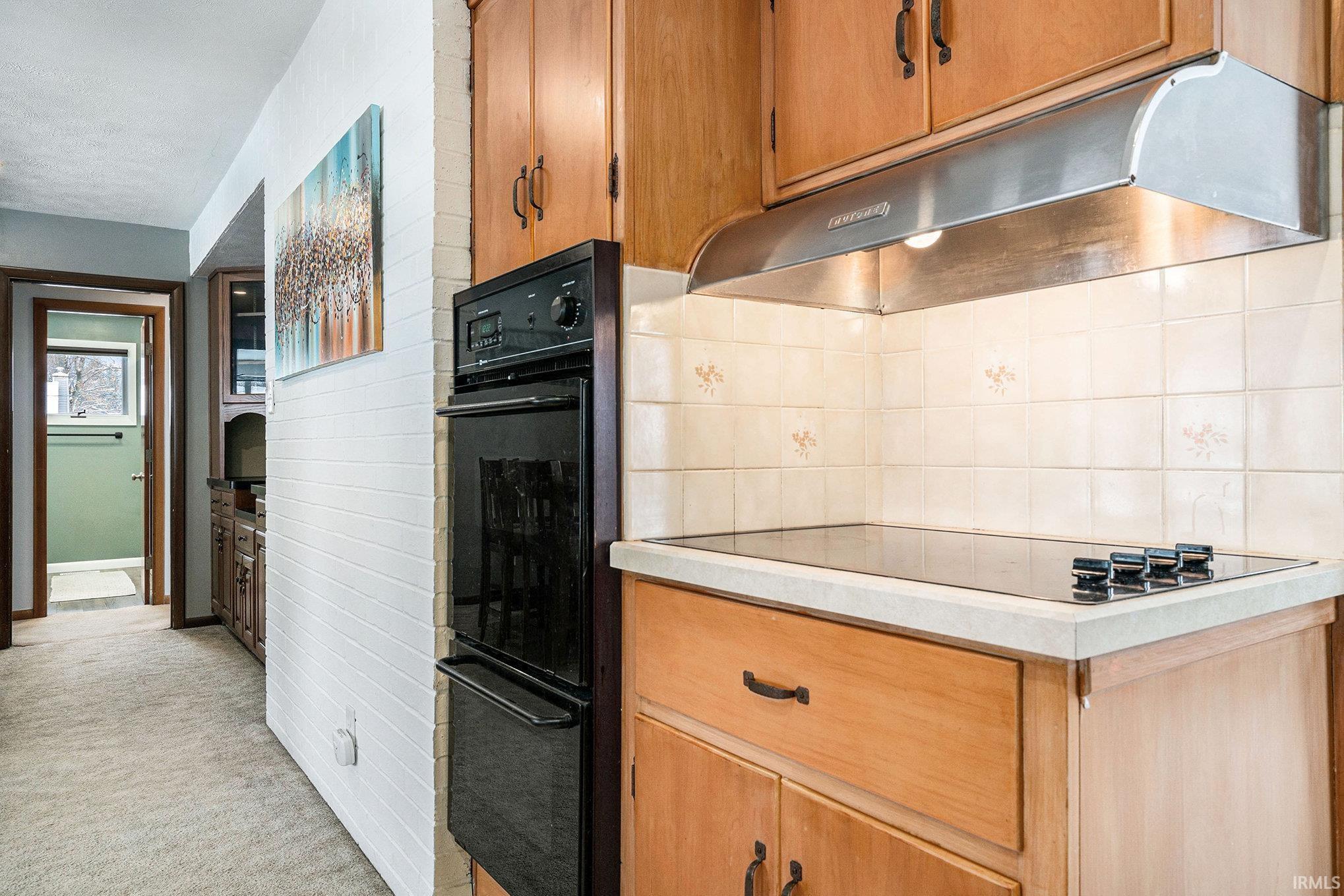 Kitchen featuring under cabinet range hood, black appliances, light countertops, light colored carpet, and backsplash