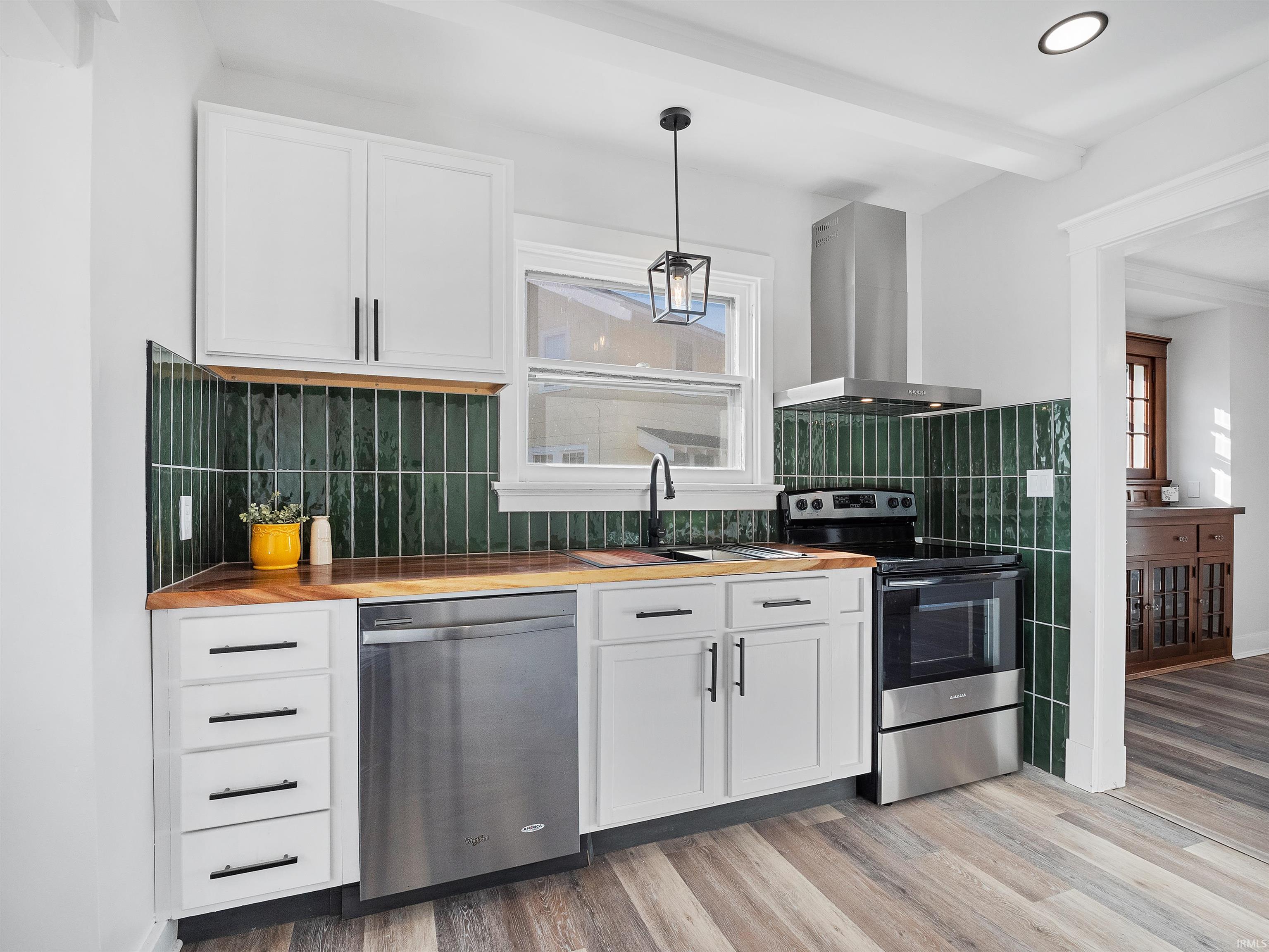 Kitchen featuring stainless steel appliances, butcher block counters, wall chimney exhaust hood, white cabinets, and beam ceiling