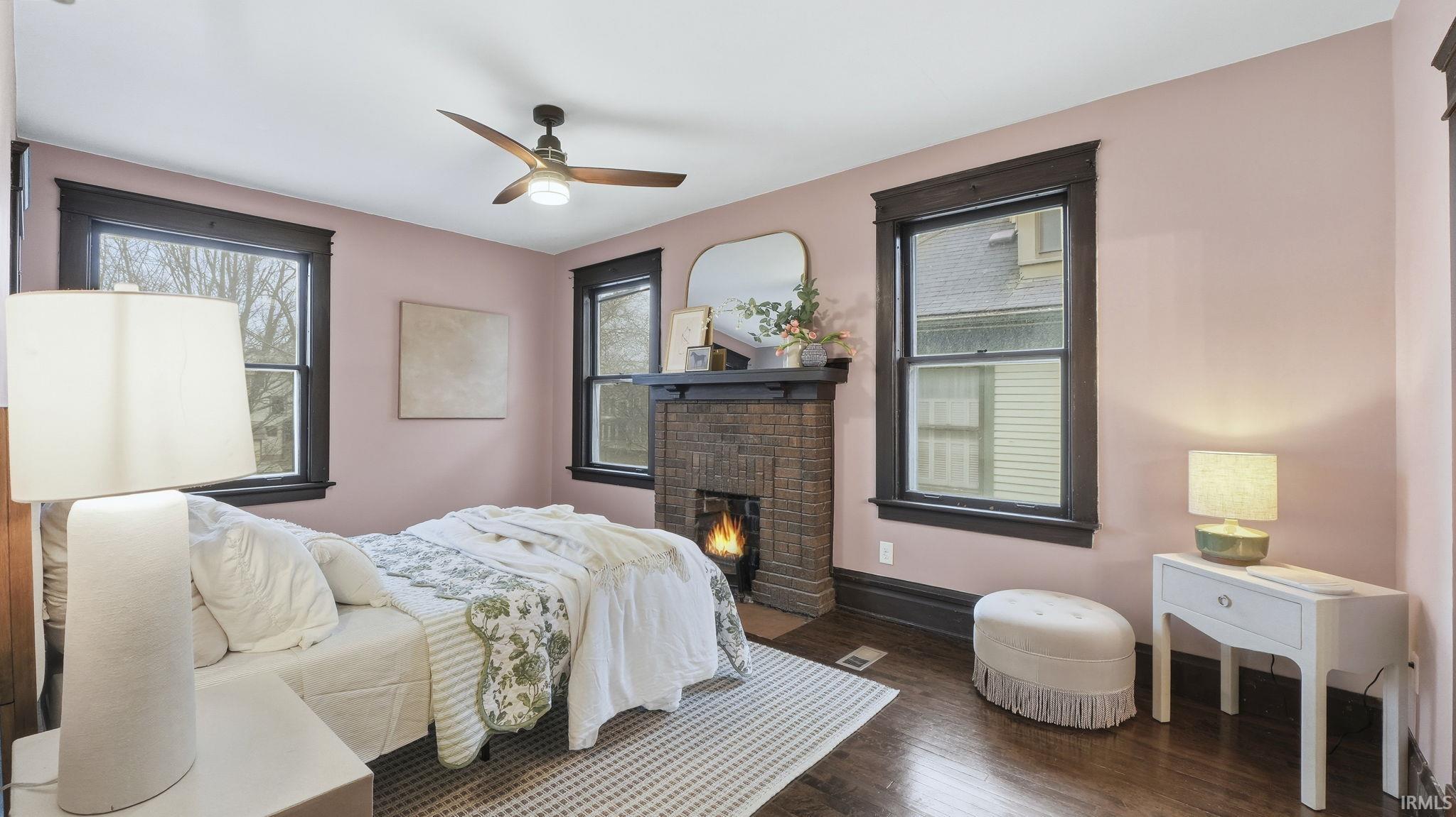 Bedroom featuring a brick fireplace, wood finished floors, a ceiling fan, and multiple windows