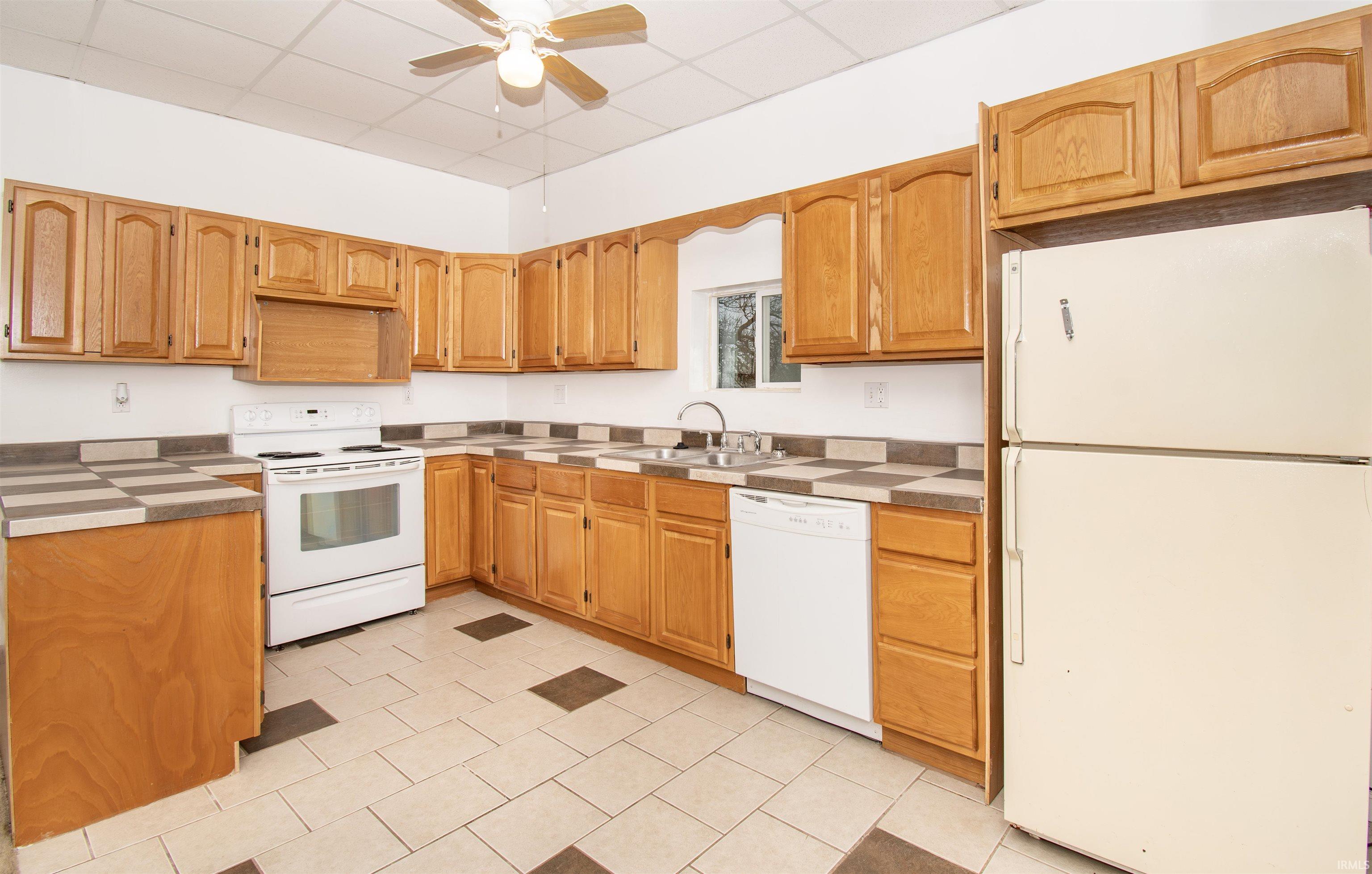 Kitchen with white appliances, ceiling fan, a drop ceiling, light tile patterned floors, and brown cabinets