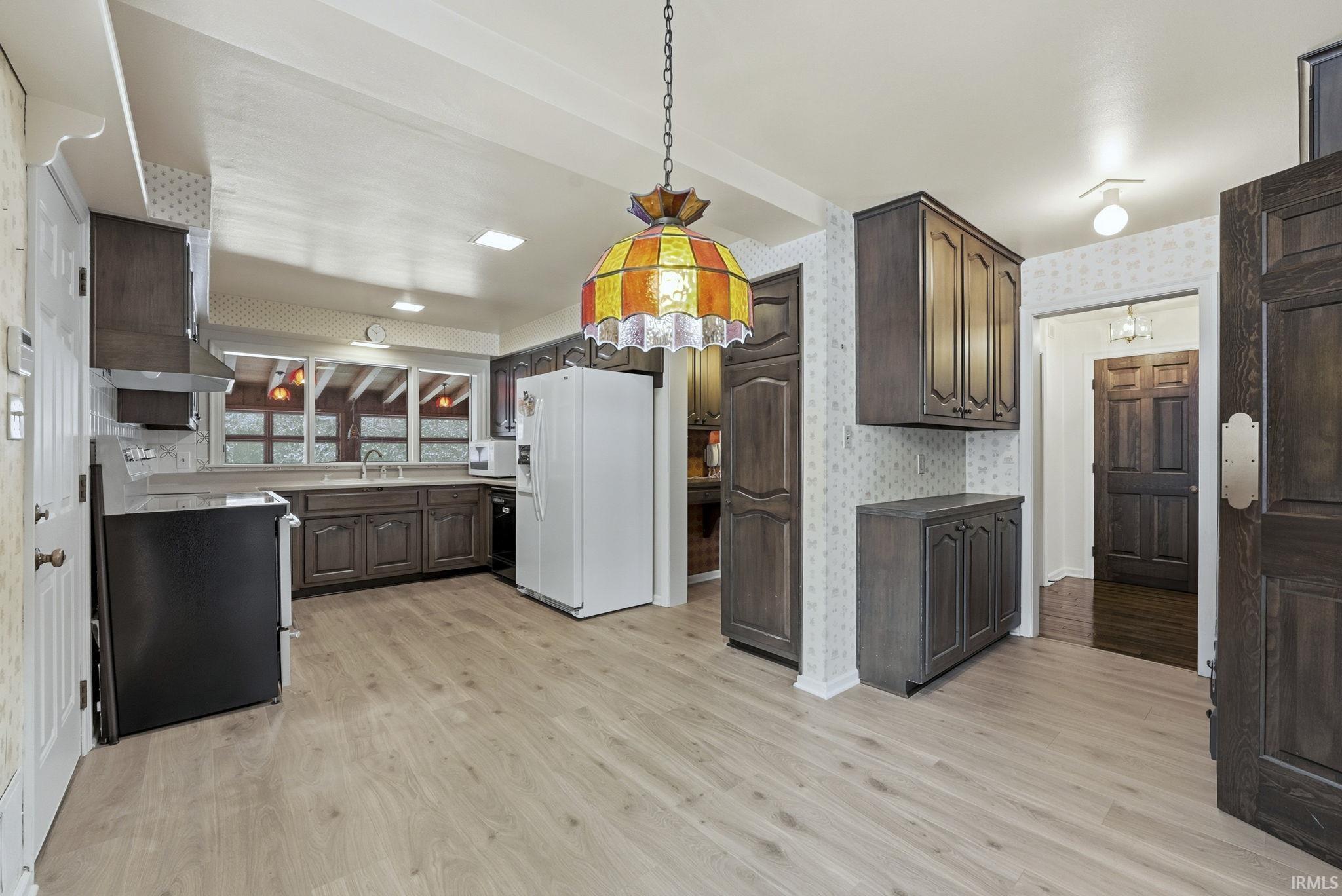 Kitchen with wallpapered walls, white appliances, hanging light fixtures, dark brown cabinets, and light wood-type flooring