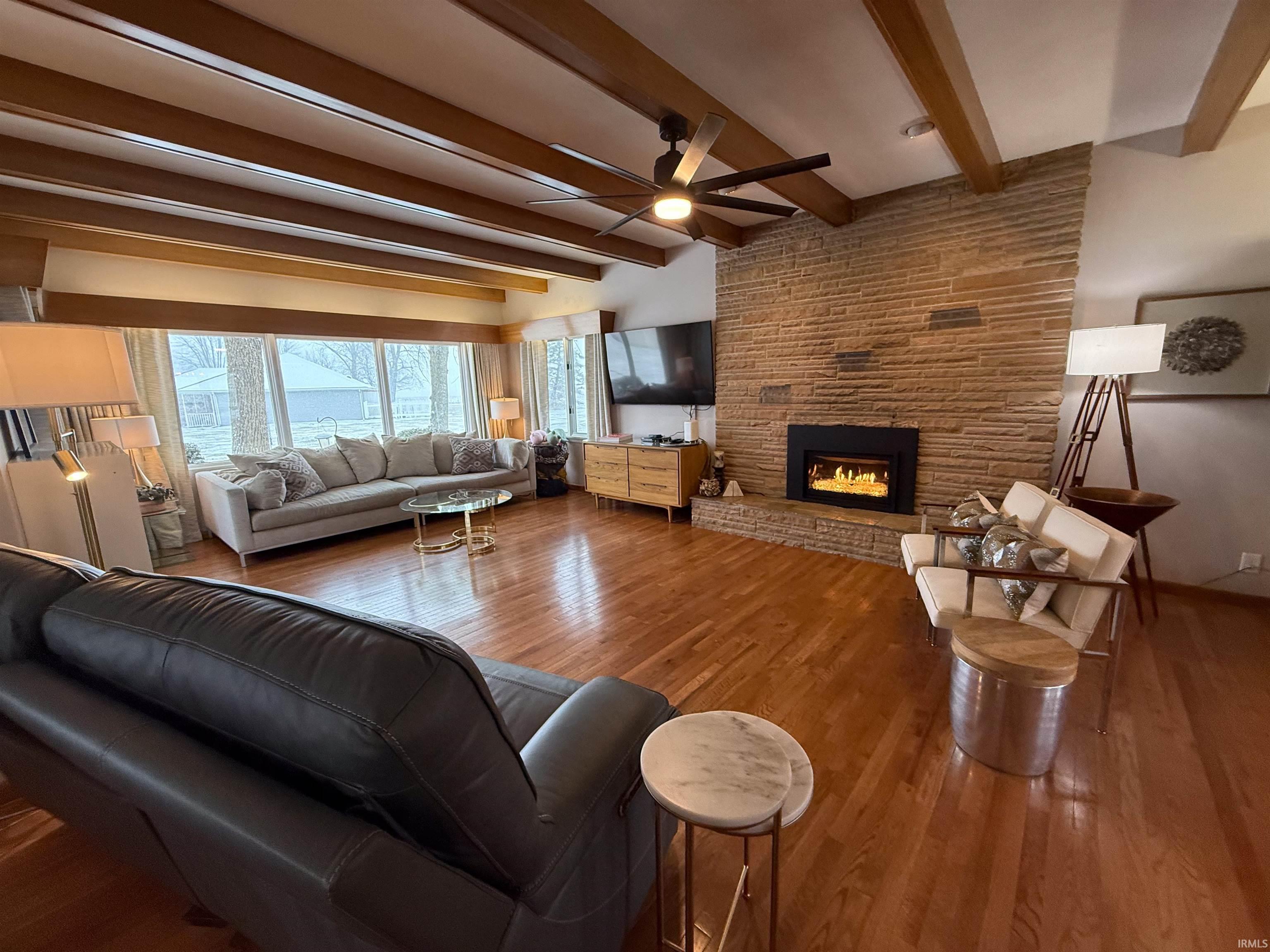 Living room featuring a large fireplace, beam ceiling, wood finished floors, and ceiling fan