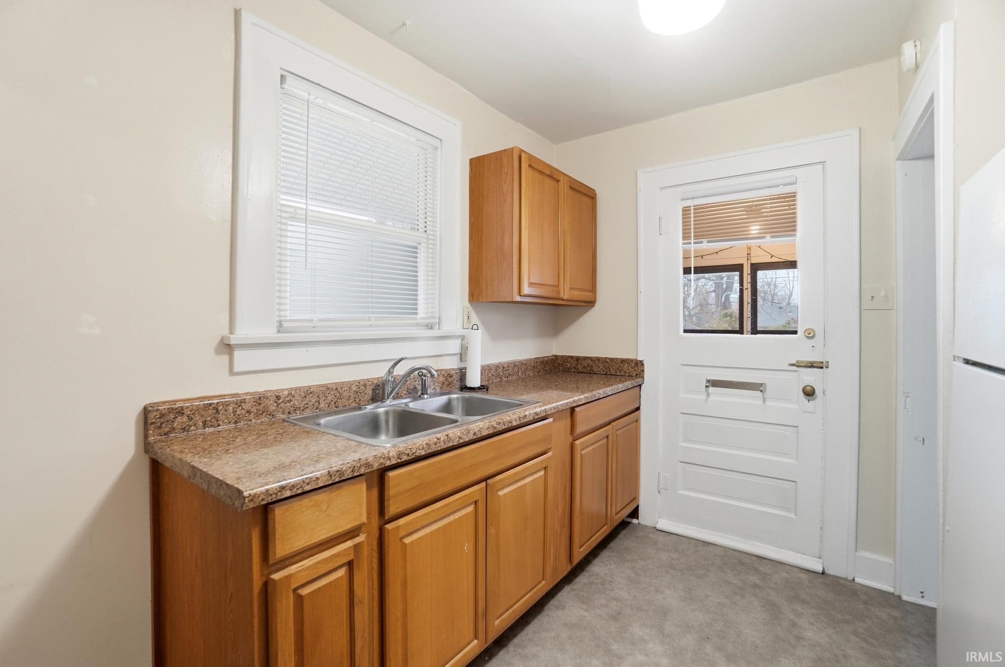 Kitchen featuring freestanding refrigerator, light carpet, brown cabinetry, and dark countertops