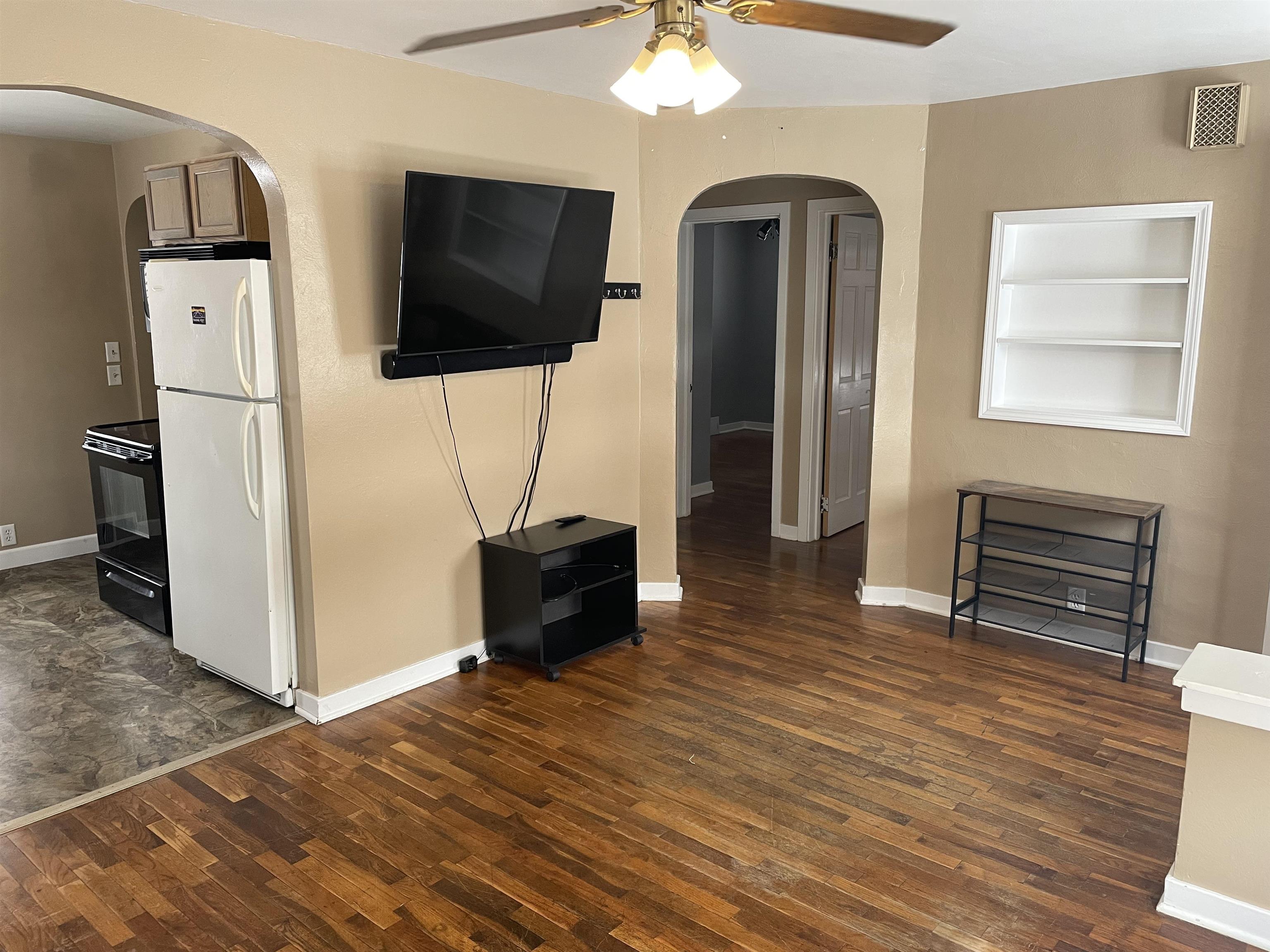 Unfurnished living room featuring arched walkways, built in shelves, dark wood-type flooring, and ceiling fan