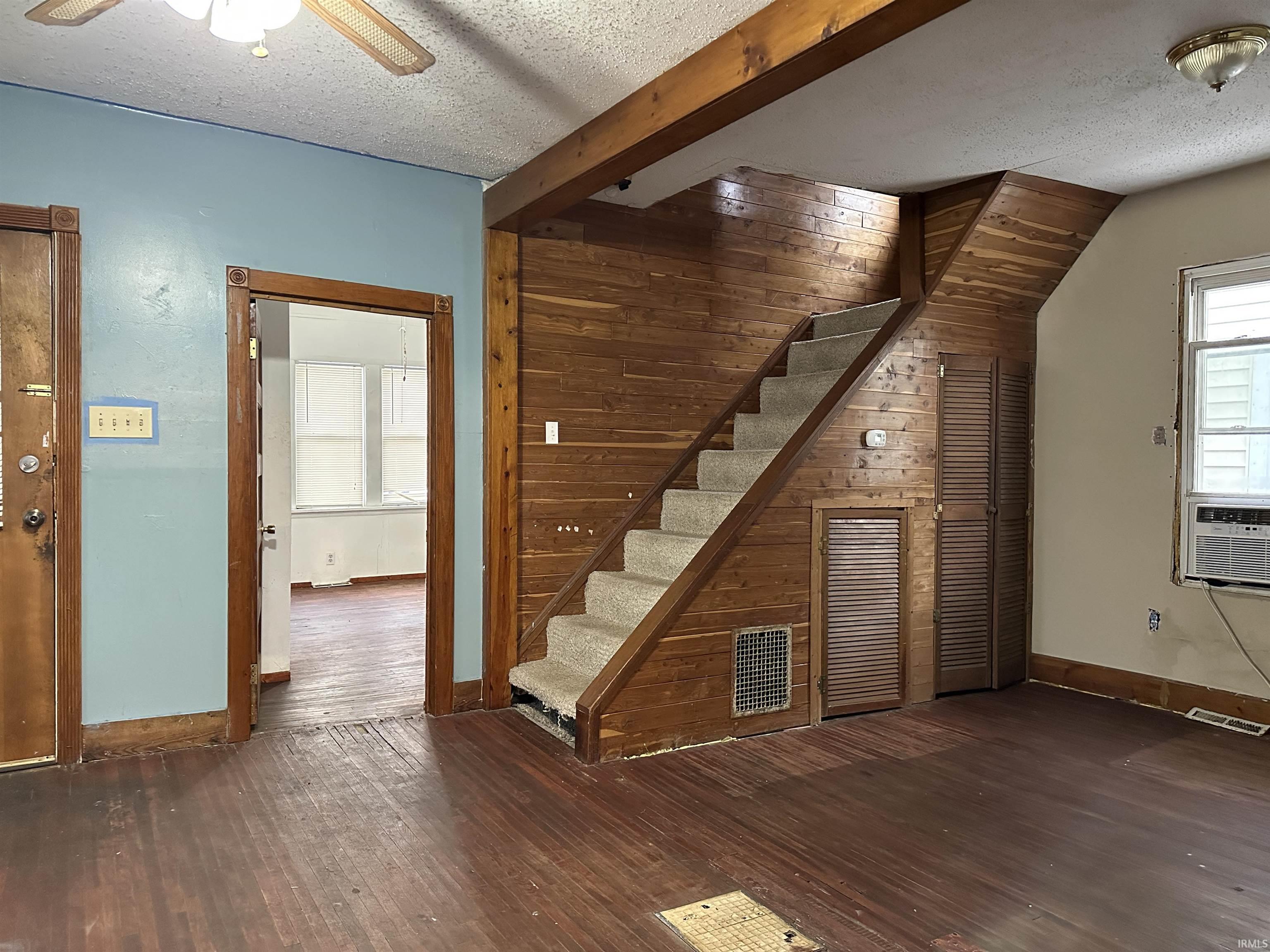 Unfurnished living room with stairway, a textured ceiling, wood walls, dark wood-style flooring, and beamed ceiling