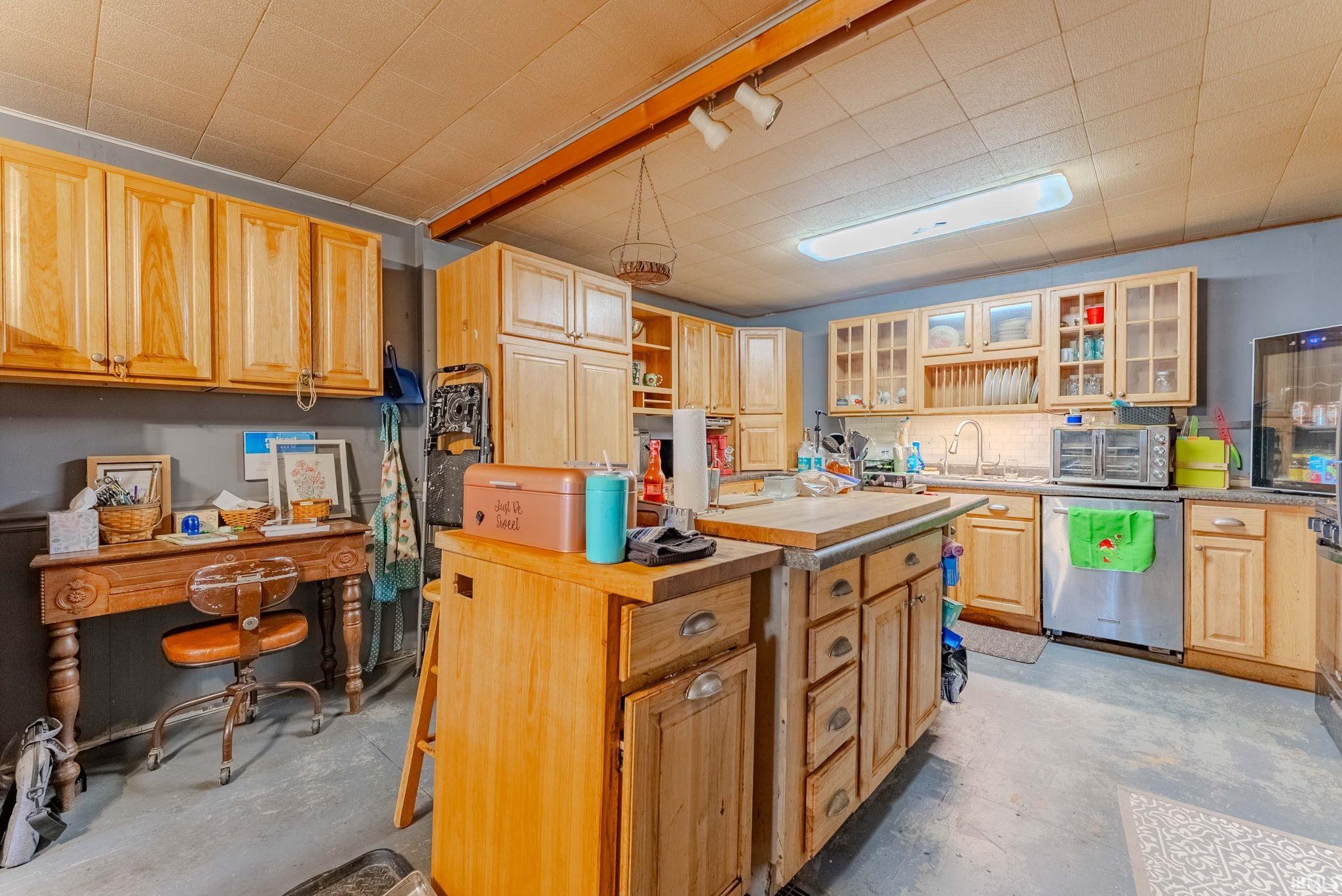 Kitchen with concrete flooring, stainless steel dishwasher, glass insert cabinets, light brown cabinets, and a kitchen island