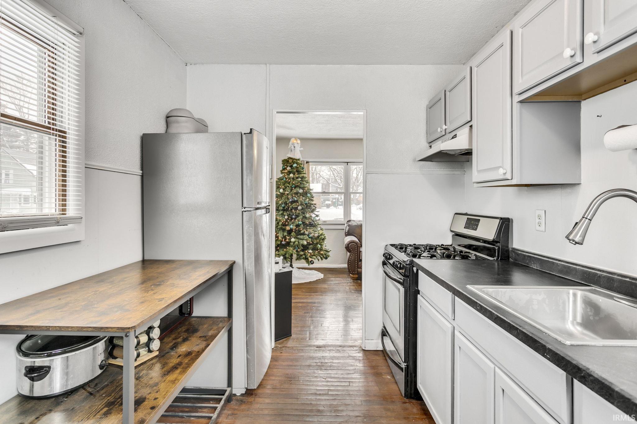 Kitchen featuring appliances with stainless steel finishes, dark countertops, under cabinet range hood, dark wood-style floors, and white cabinets