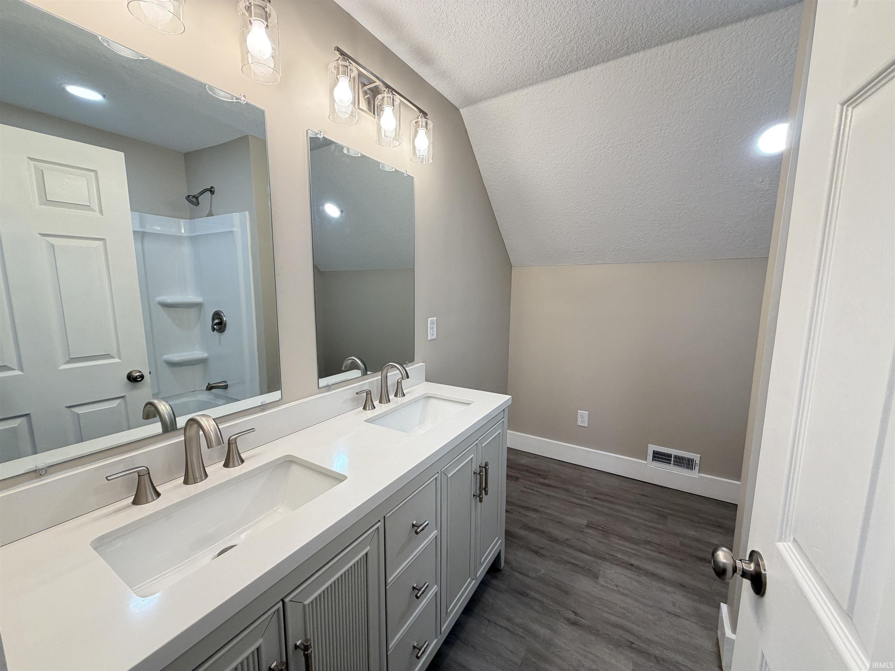 Bathroom featuring a textured ceiling, double vanity, vaulted ceiling, dark wood-style flooring, and  shower combination