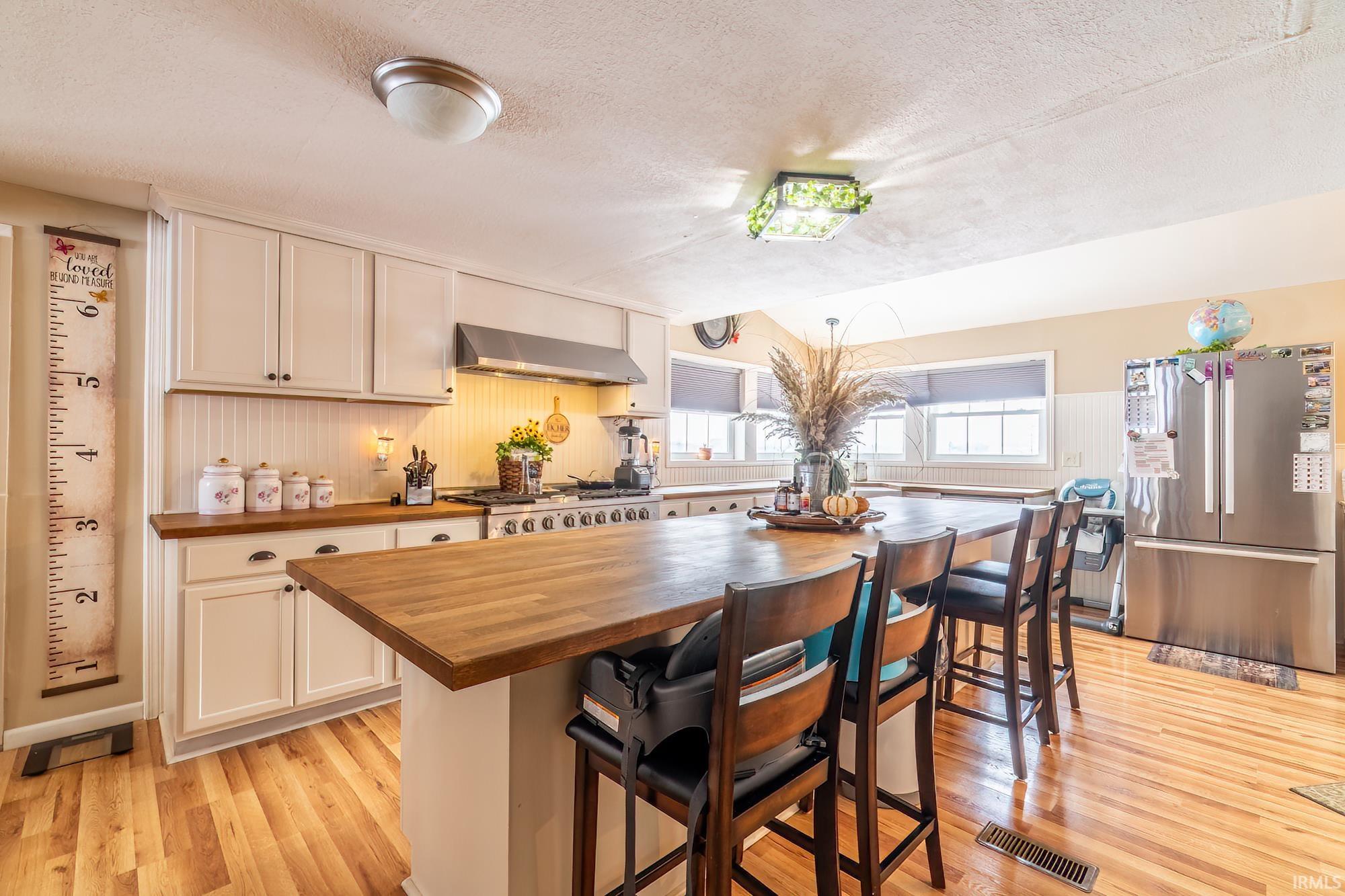 Kitchen featuring a kitchen bar, freestanding refrigerator, wall chimney range hood, light wood finished floors, and a textured ceiling