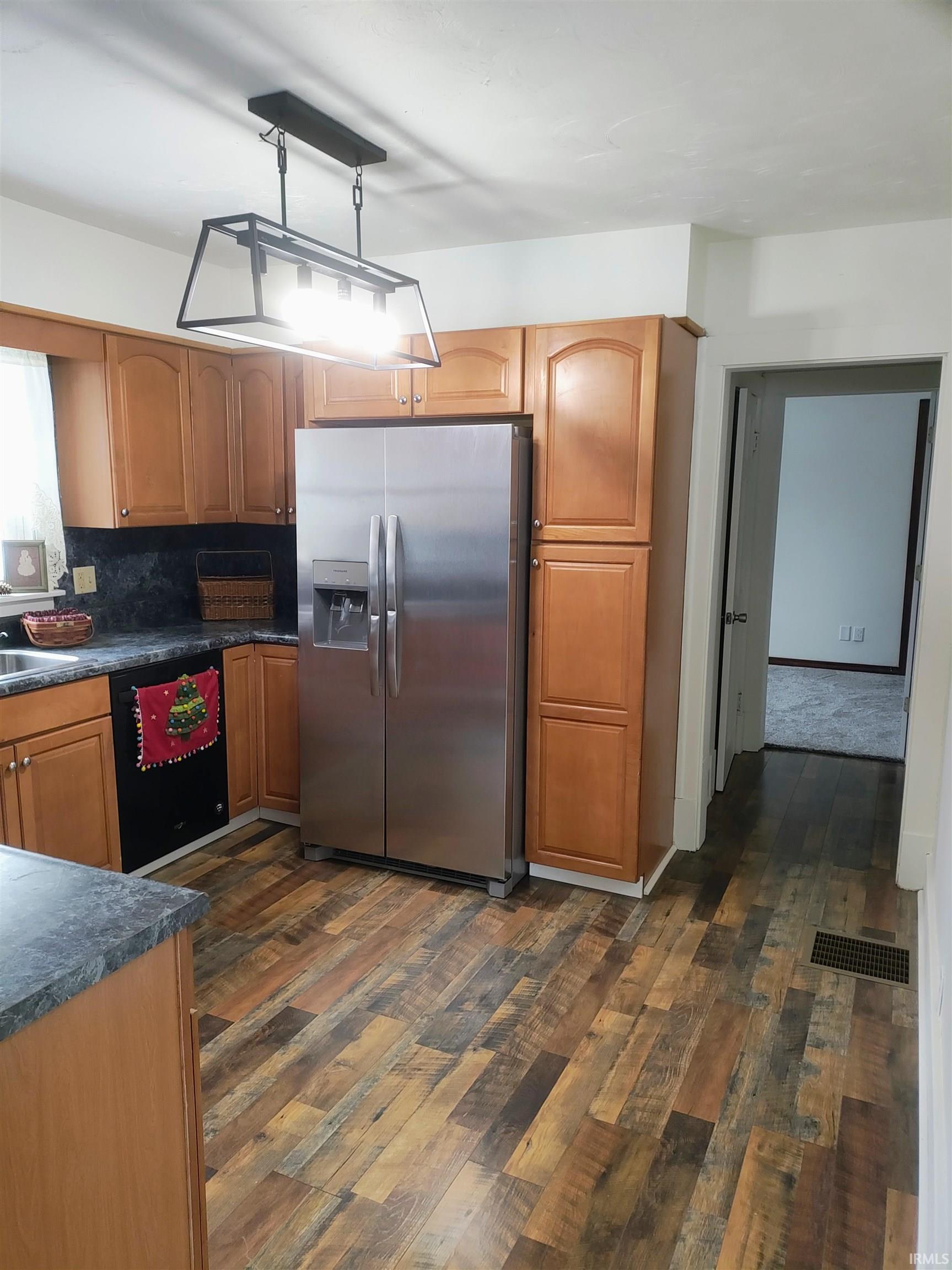 Kitchen with dark wood finished floors, stainless steel fridge, brown cabinetry, dishwasher, and dark countertops
