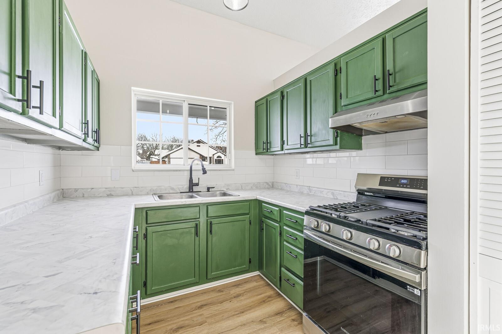 Kitchen with green cabinets, stainless steel range with gas stovetop, light countertops, under cabinet range hood, and backsplash