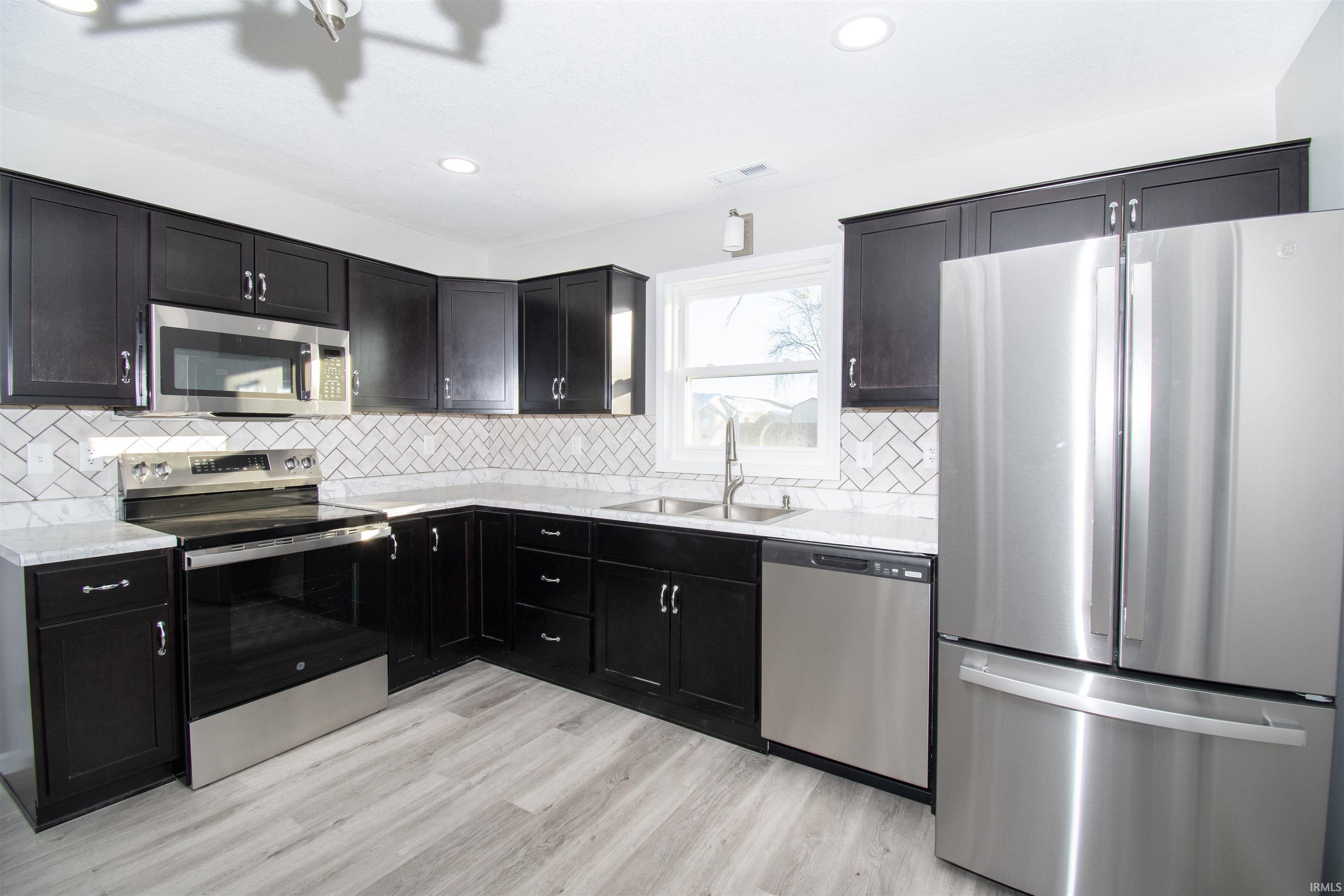 Kitchen featuring stainless steel appliances, dark cabinets, light stone counters, backsplash, and recessed lighting