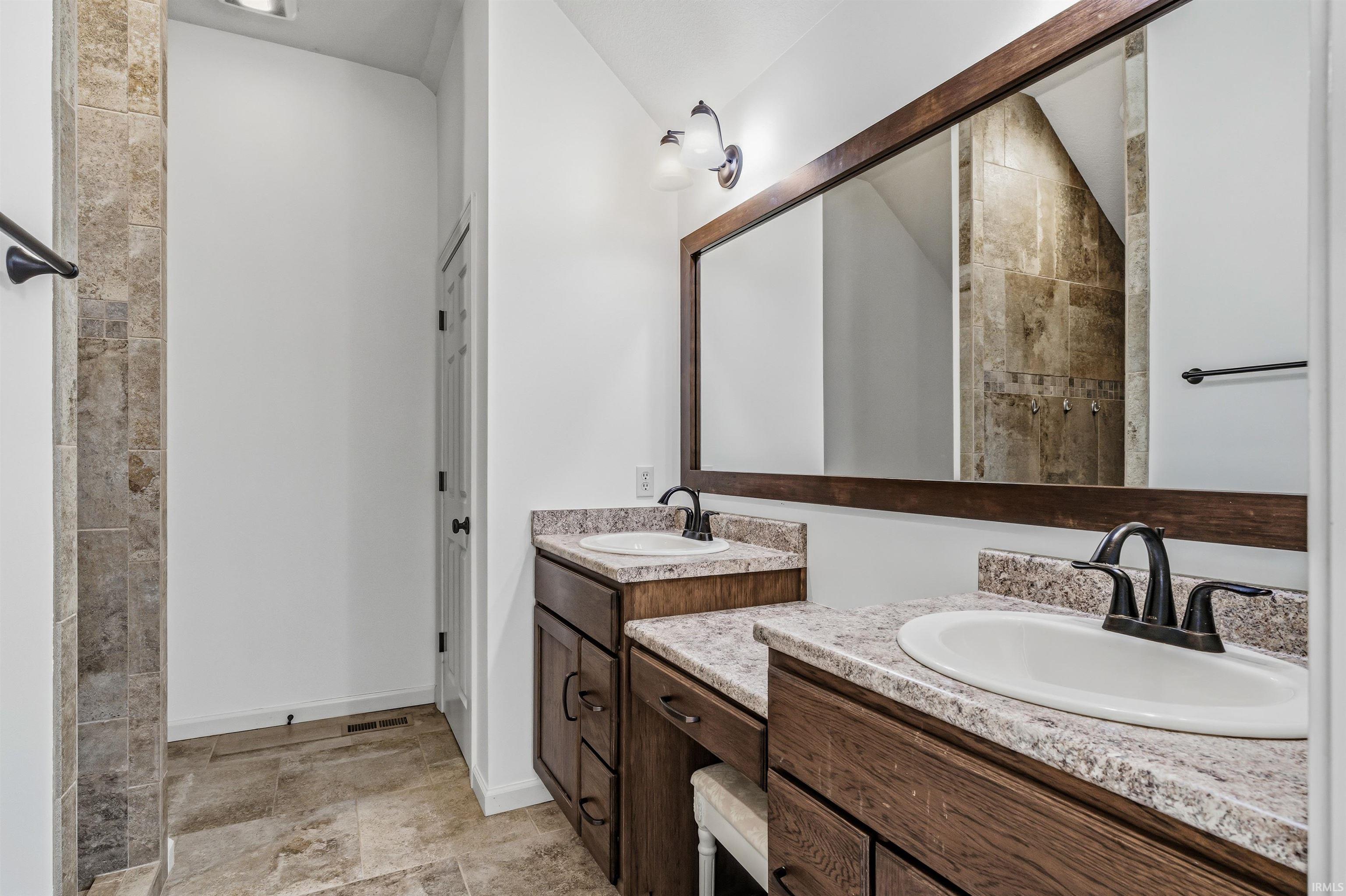 Bathroom featuring two vanities, vaulted ceiling, a tile shower, and stone tile flooring