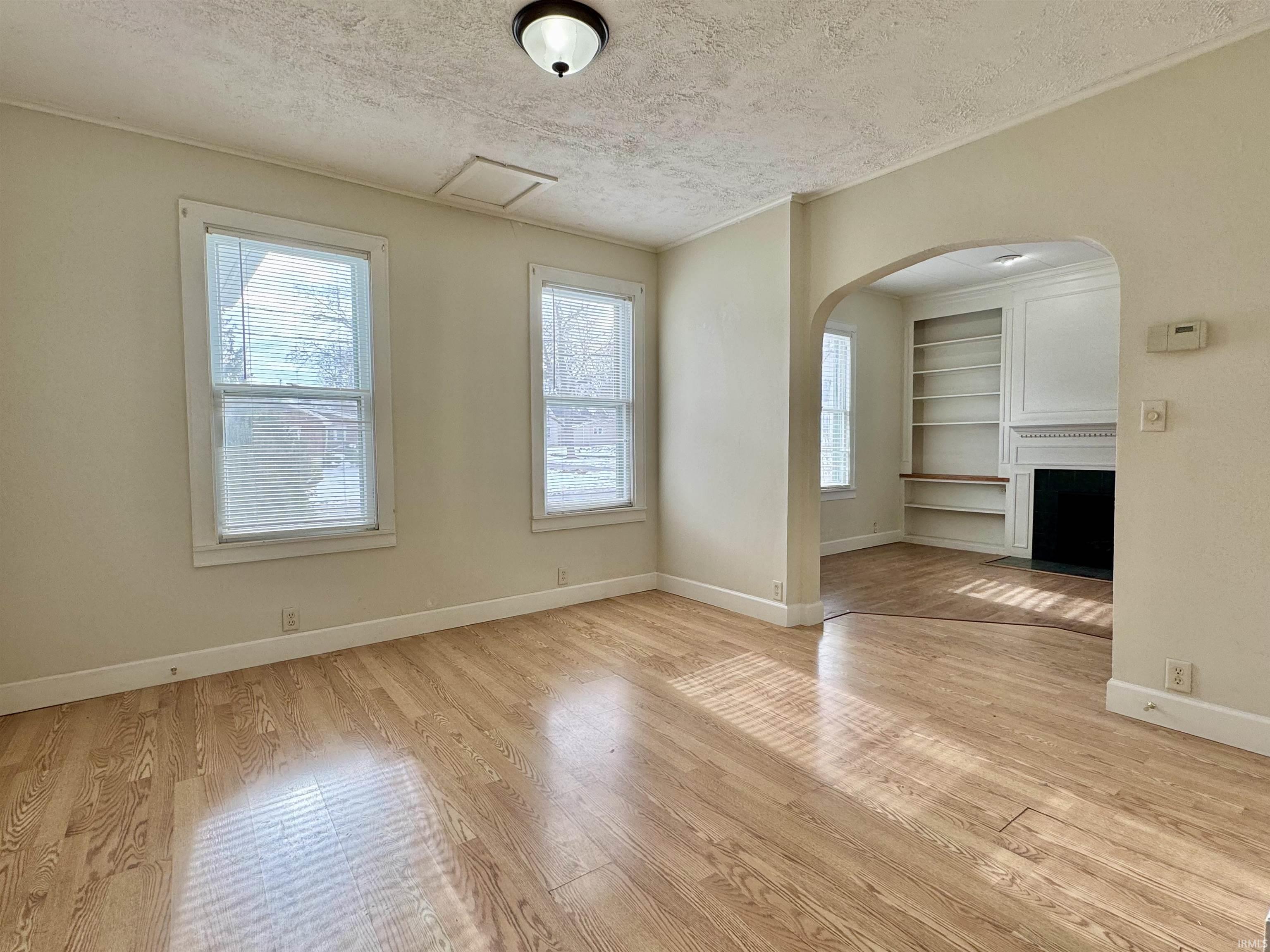 Unfurnished living room featuring a textured ceiling, light wood-type flooring, built in shelves, a fireplace with flush hearth, and arched walkways