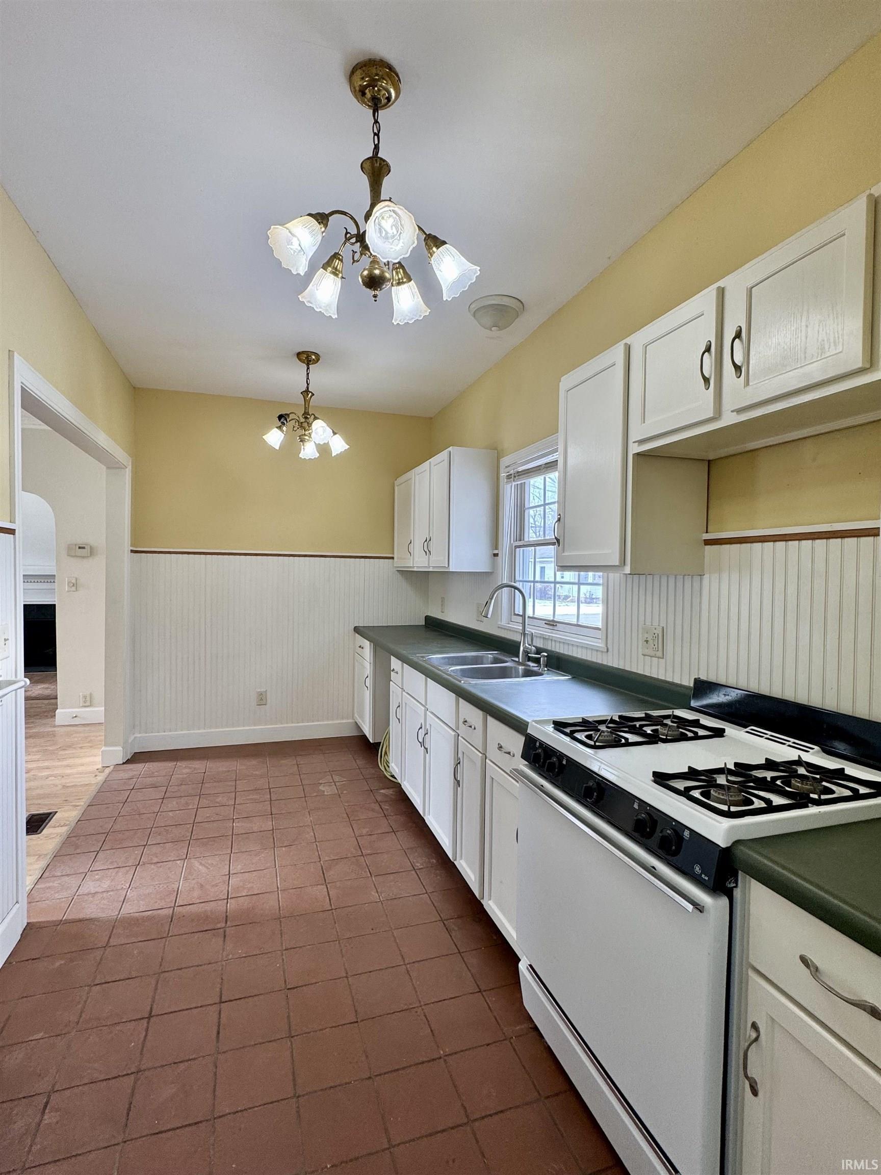 Kitchen featuring white range with gas stovetop, wainscoting, white cabinets, dark countertops, and hanging light fixtures