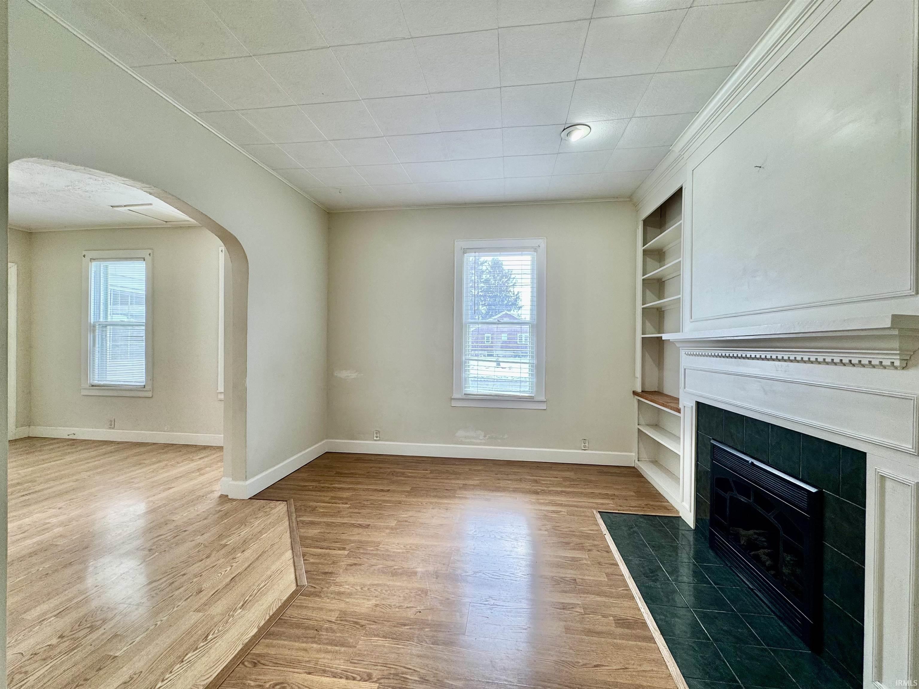 Unfurnished living room featuring arched walkways, built in shelves, a tile fireplace, light wood finished floors, and crown molding
