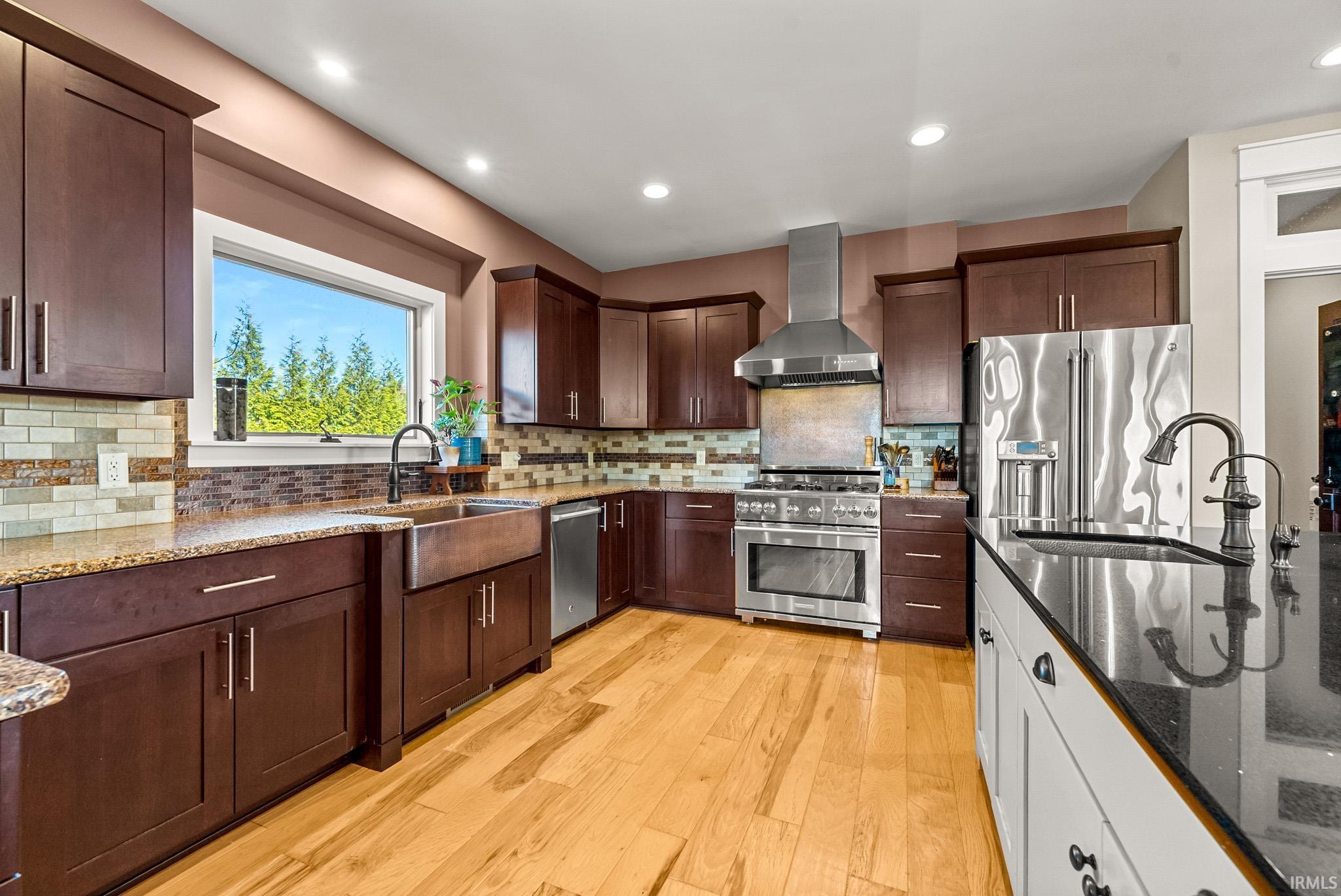 Kitchen featuring dark stone counters, stainless steel appliances, wall chimney range hood, dark brown cabinetry, and recessed lighting