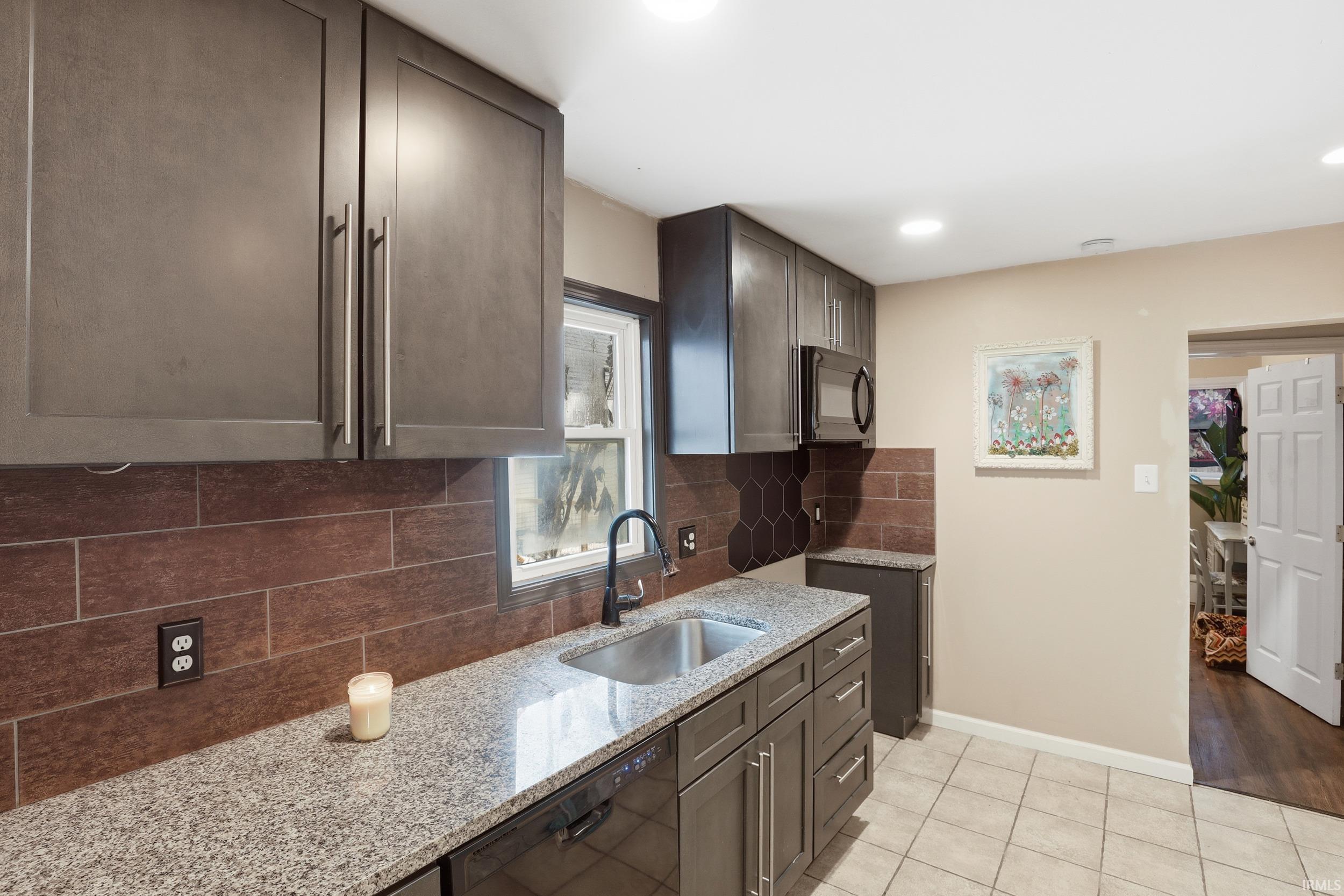 Kitchen featuring light stone countertops, backsplash, black appliances, light tile patterned floors, and dark brown cabinetry