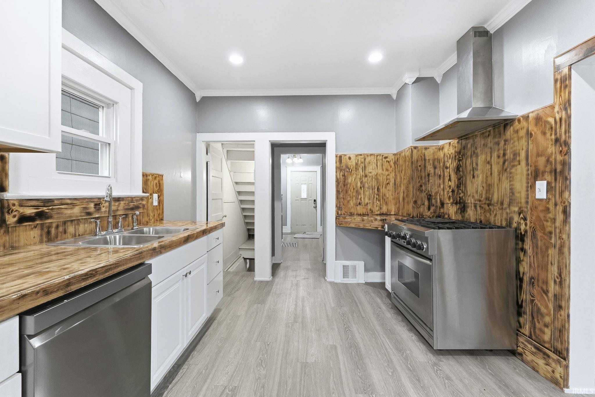 Kitchen featuring appliances with stainless steel finishes, crown molding, wall chimney range hood, white cabinets, and light wood-style floors