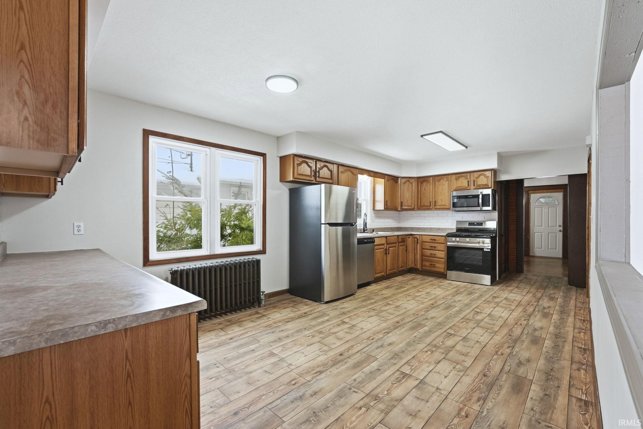 Kitchen featuring brown cabinets, appliances with stainless steel finishes, radiator heating unit, light countertops, and light wood-style flooring