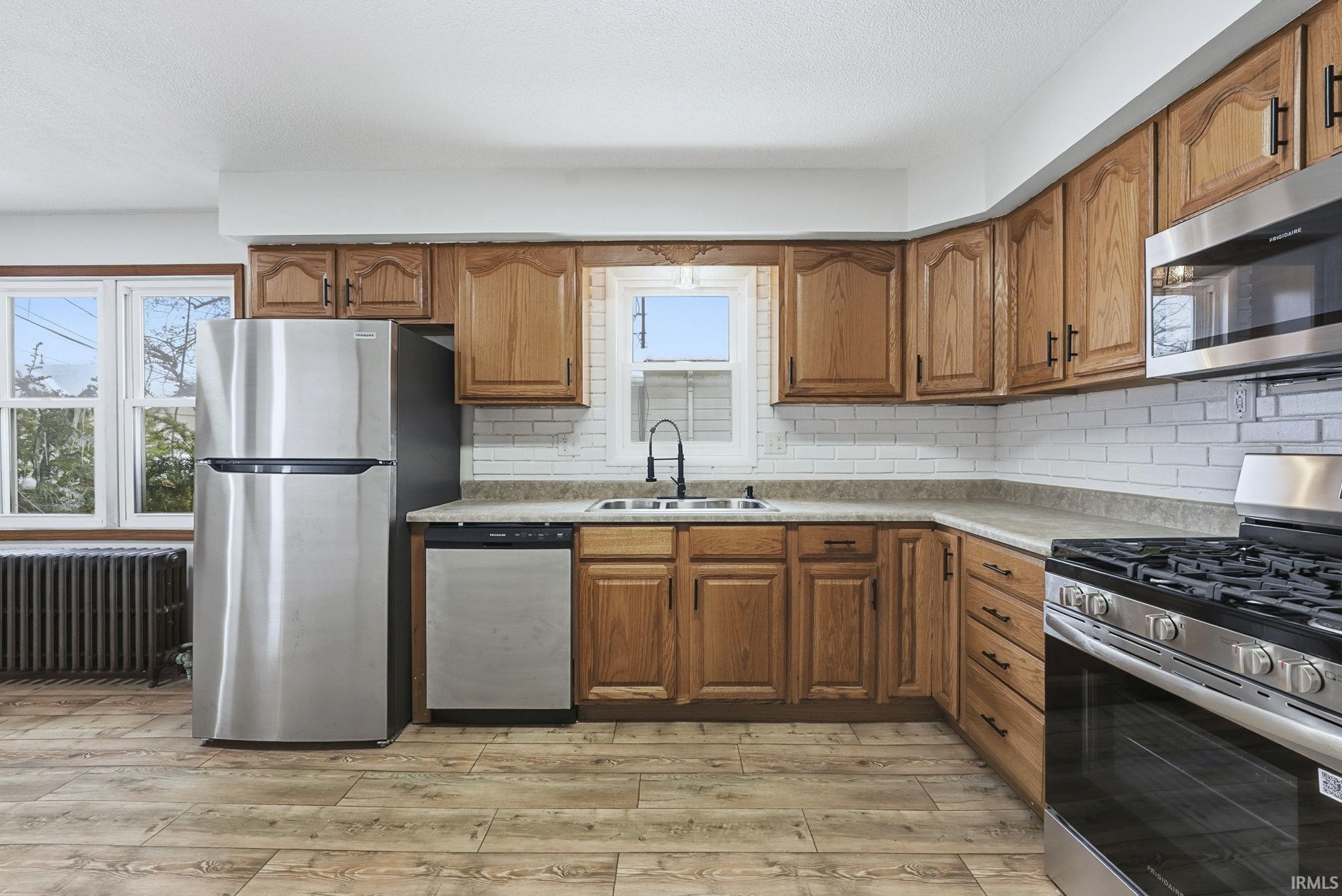 Kitchen with brown cabinetry, appliances with stainless steel finishes, radiator, light countertops, and light wood-style floors