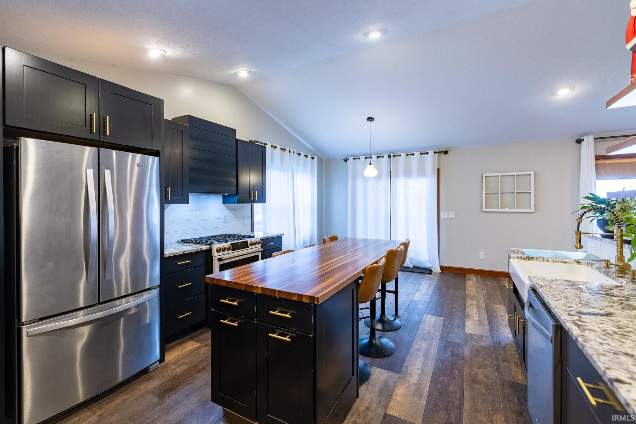 Kitchen featuring appliances with stainless steel finishes, dark wood-style flooring, dark stone countertops, decorative light fixtures, and vaulted ceiling