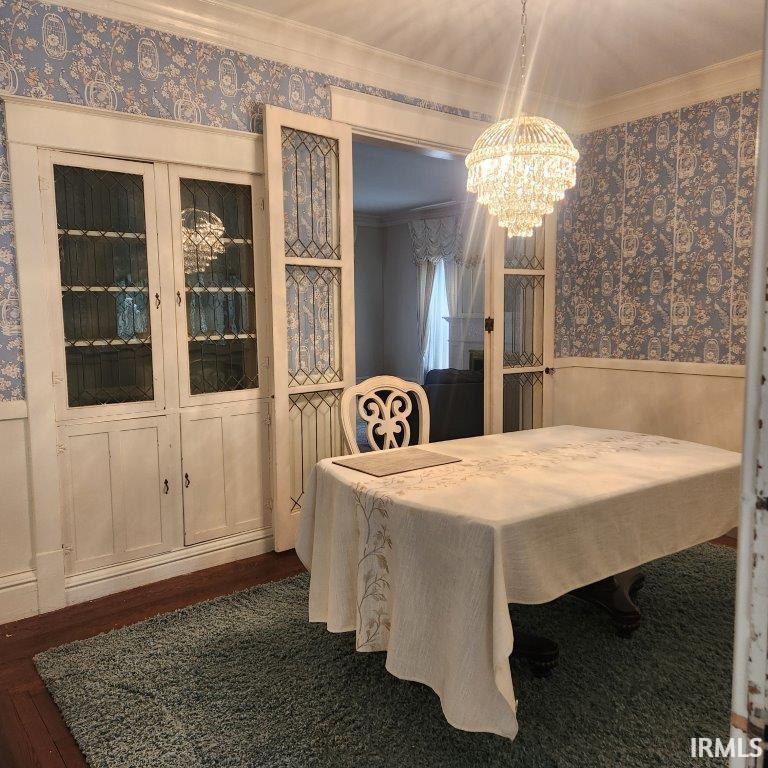 Dining space featuring wallpapered walls, wainscoting, ornamental molding, dark wood-style floors, and a chandelier