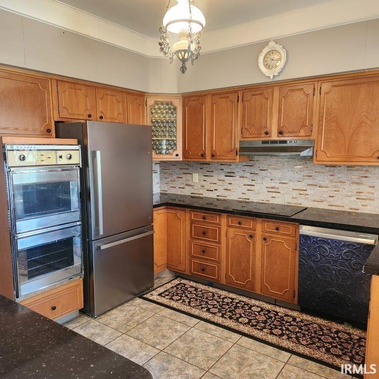 Kitchen with appliances with stainless steel finishes, brown cabinetry, backsplash, dark stone counters, and under cabinet range hood
