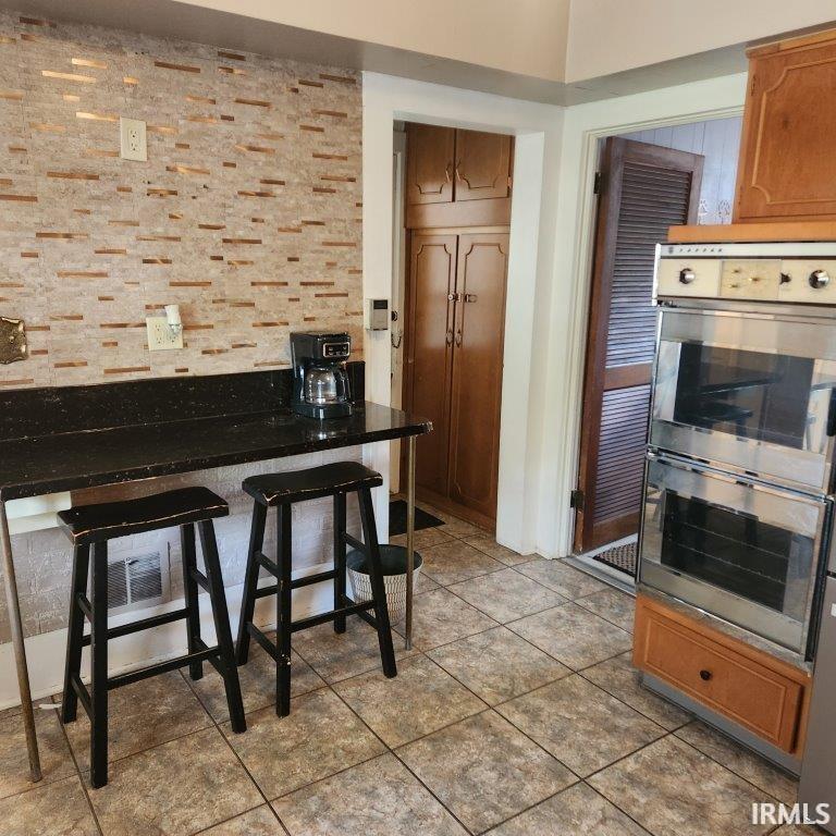 Kitchen with stainless steel double oven, dark countertops, brown cabinets, and a breakfast bar