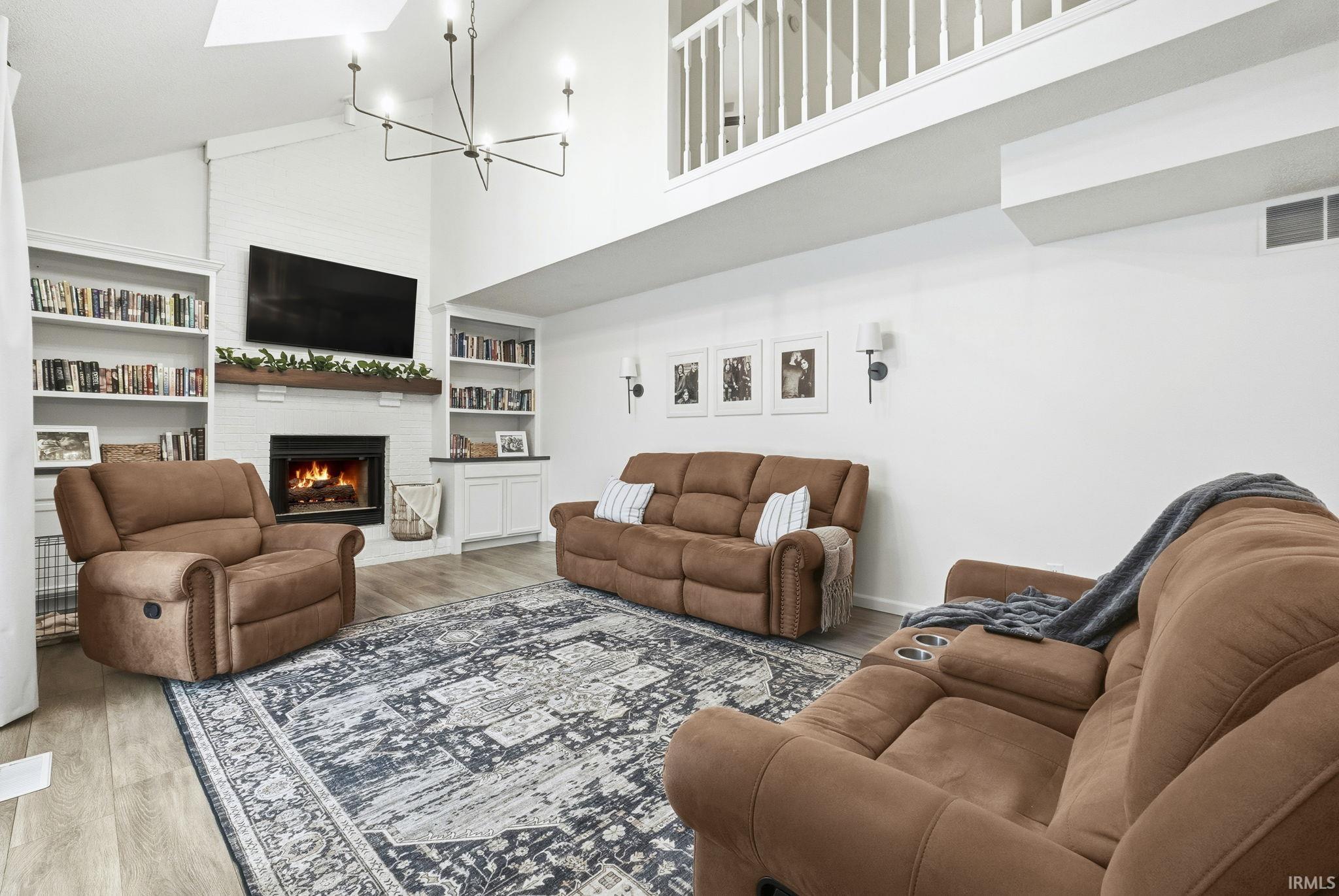 Living room featuring high vaulted ceiling, wood finished floors, a brick fireplace, built in features, and a chandelier