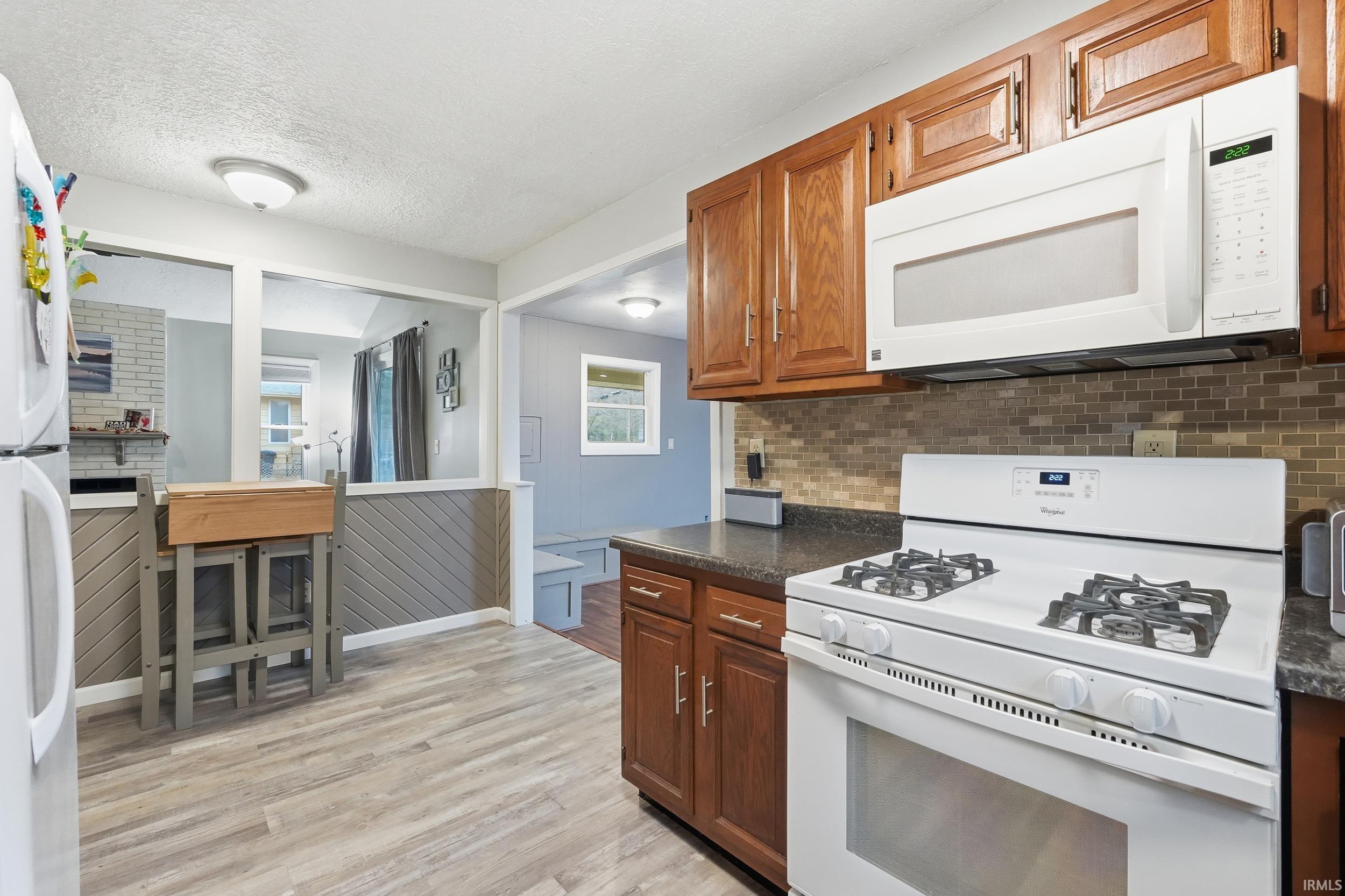 Kitchen featuring white appliances, dark countertops, light wood-type flooring, a textured ceiling, and tasteful backsplash